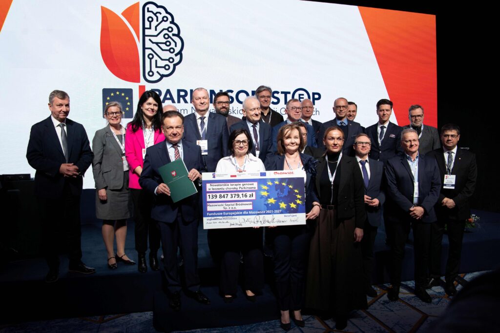 Grupa formalnie ubranych osób stoi na scenie, trzymając duży uroczysty czek i flagę UE. Pozują do zdjęcia przed tłem z cyfrowym logo mózgu i napisem "Parkinson STOP". Większość z nich uśmiecha się i patrzy wobiektyw.