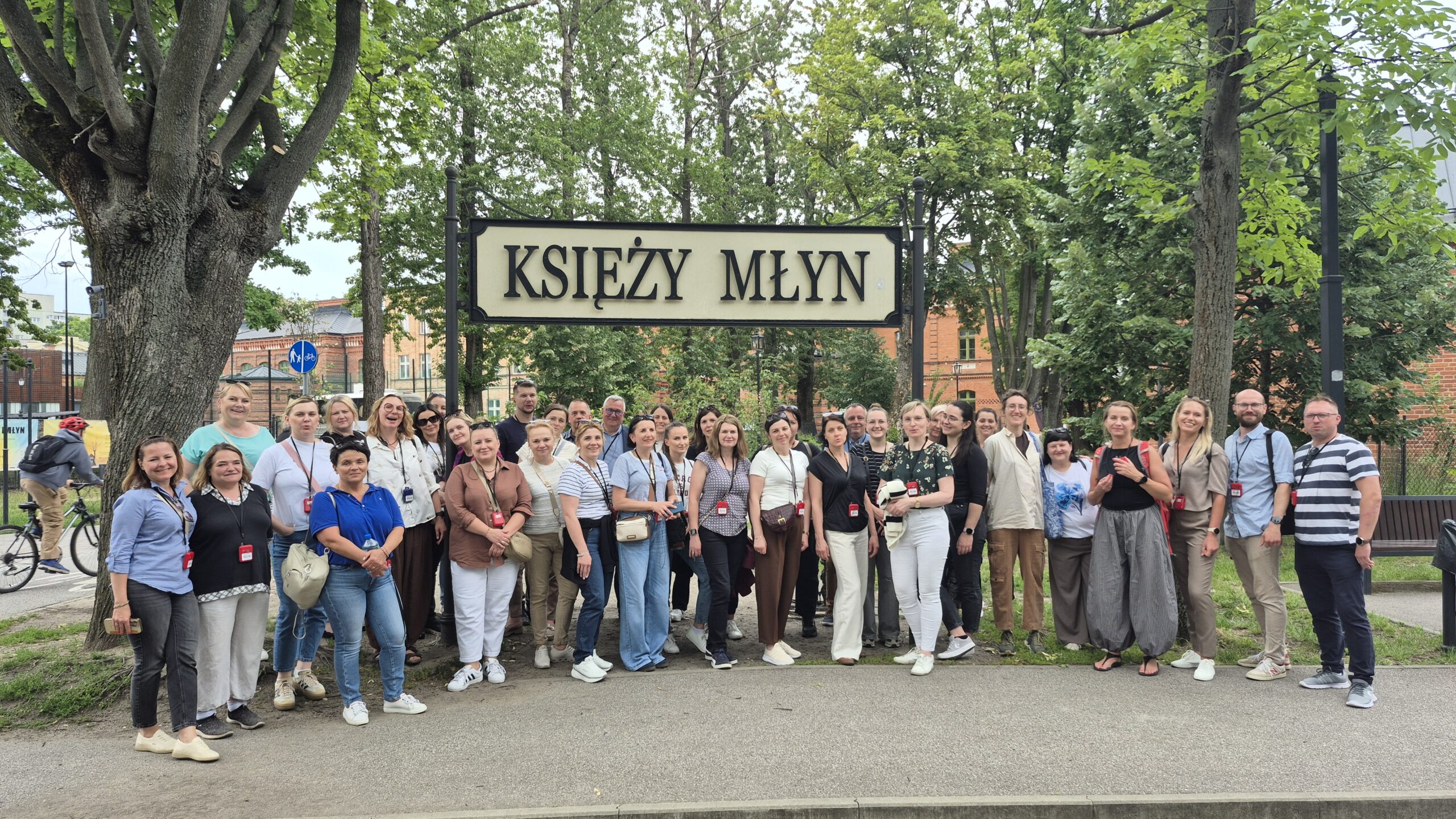 Duża grupa ludzi pozuje na świeżym powietrzu przed tablicą z napisem „KSIĘŻY MŁYN”. Stoją na chodniku otoczonym drzewami, a w tle widać kilka budynków. Większość osób się uśmiecha, a pogoda wydaje się łagodna i pochmurna.