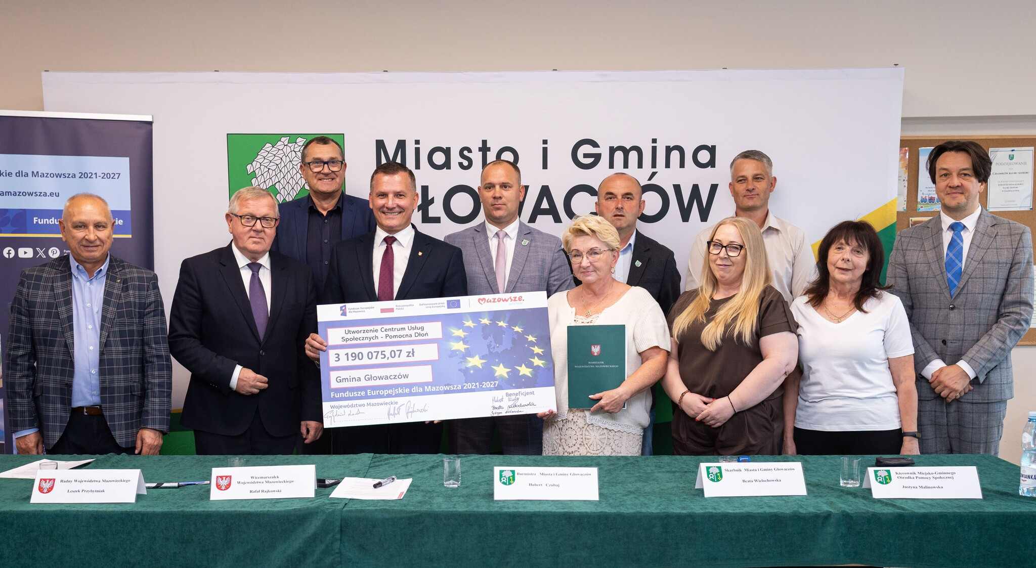 Grupa dwunastu osób w strojach formalnych stoi w rzędzie w sali konferencyjnej przed banerem z napisem „Miasto i Gmina GŁOWACZÓW”, na środku mężczyzna w garniturze trzyma duży symboliczny czek z motywem gwiazd Unii Europejskiej i widoczną kwotą 3 190 075,07 zł, obok kobieta trzyma zieloną teczkę, a przed nimi długi stół z zielonym obrusem i imiennymi karteczkami.