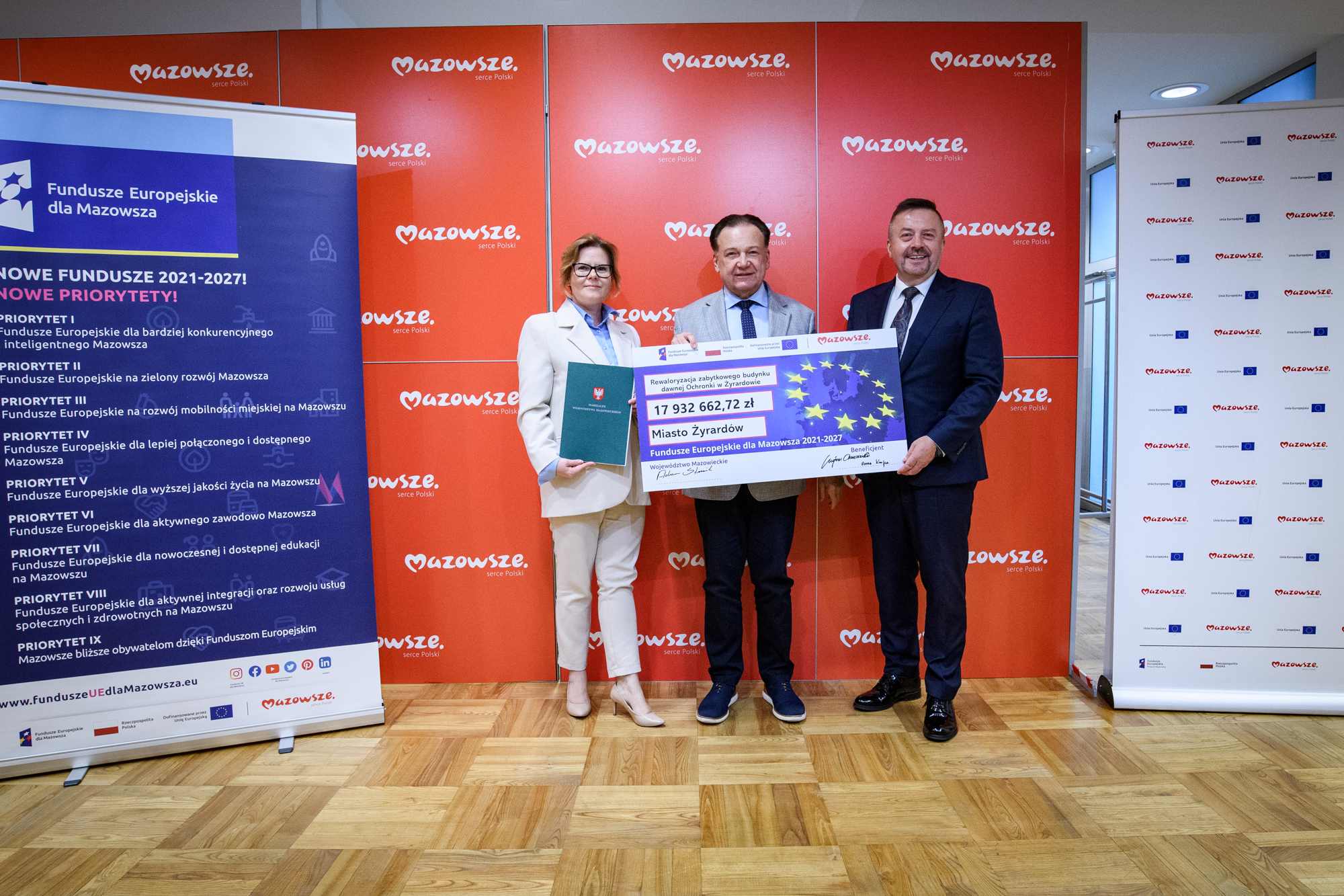 Trzy osoby w strojach formalnych stoją na środku przed czerwonym panelem z powtarzającym się logo „Mazowsze”, trzymając duży symboliczny czek z gwiazdami UE, napisem „Miasto Żyrardów” i kwotą 17 932 662,72 zł; po lewej widoczny niebieski banner „Fundusze Europejskie dla Mazowsza”, po prawej panel z małymi logotypami, a całość na drewnianej podłodze.