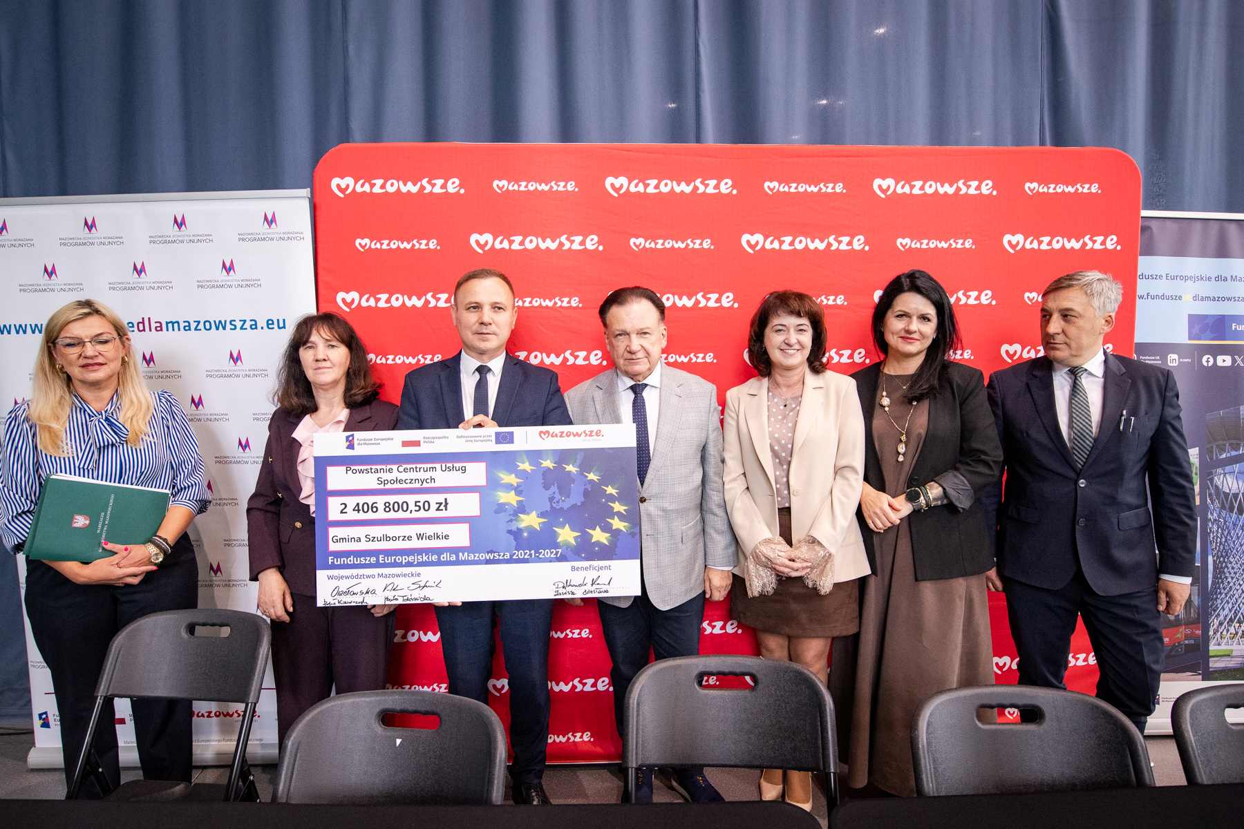 Siedmioro osób w formalnych strojach stoi przed czerwonym banerem z powtarzającym się napisem "Mazowsze", trzymając duży symboliczny czek na 2 406 800,50 zł z napisem "Powstanie Centrum Usług Społecznych" i "Gmina Szulborze Wielkie", po bokach widoczne roll-upy informacyjne, a na pierwszym planie kilka pustych czarnych krzeseł.