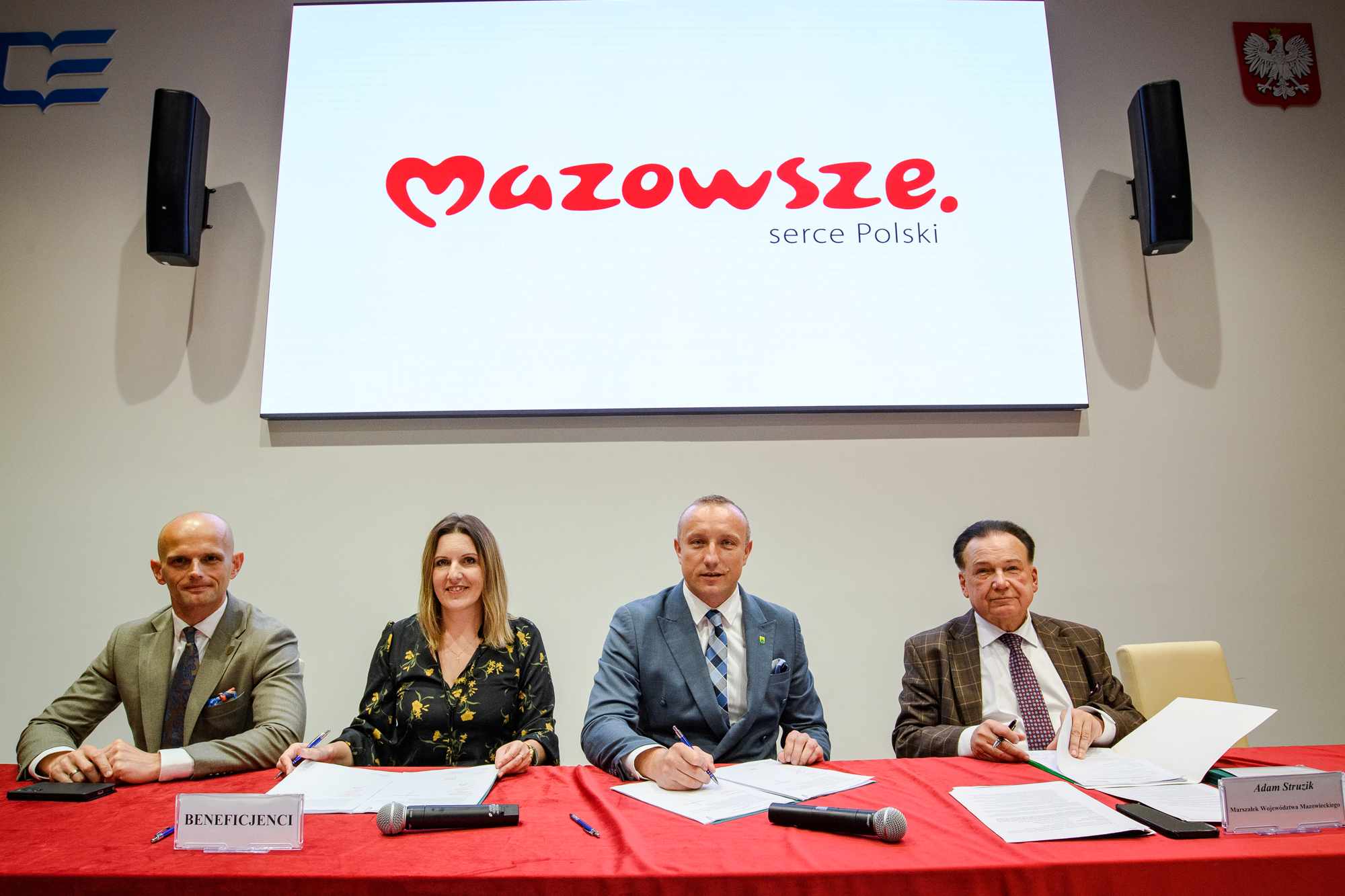 Cztery osoby siedzą przy stole z czerwoną narzutą podpisując dokumenty, nad nimi duży ekran z czerwonym logotypem „Mazowsze serce Polski”, na stole mikrofony, stosy papierów i tabliczka z napisem „BENEFICJENCI”, a po bokach głośniki i godło Polski na ścianie.