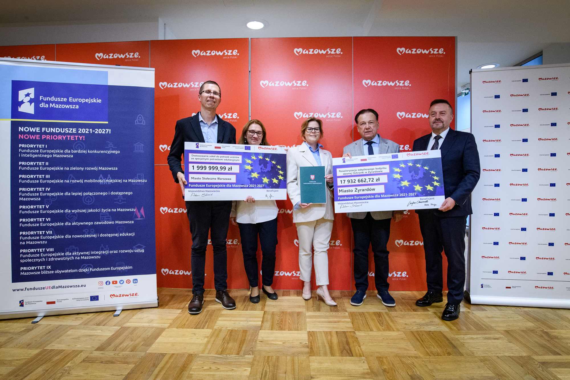 Pięć osób stojących w rzędzie na drewnianej podłodze przed czerwonym panelem z powtarzającym się logo „Mazowsze”, dwie osoby trzymają duże symboliczne czeki z widocznymi kwotami 1 999 999,99 zł i 17 932 662,72 zł, jedna osoba trzyma zielony folder, a po bokach umieszczone są pionowe banery informacyjne.