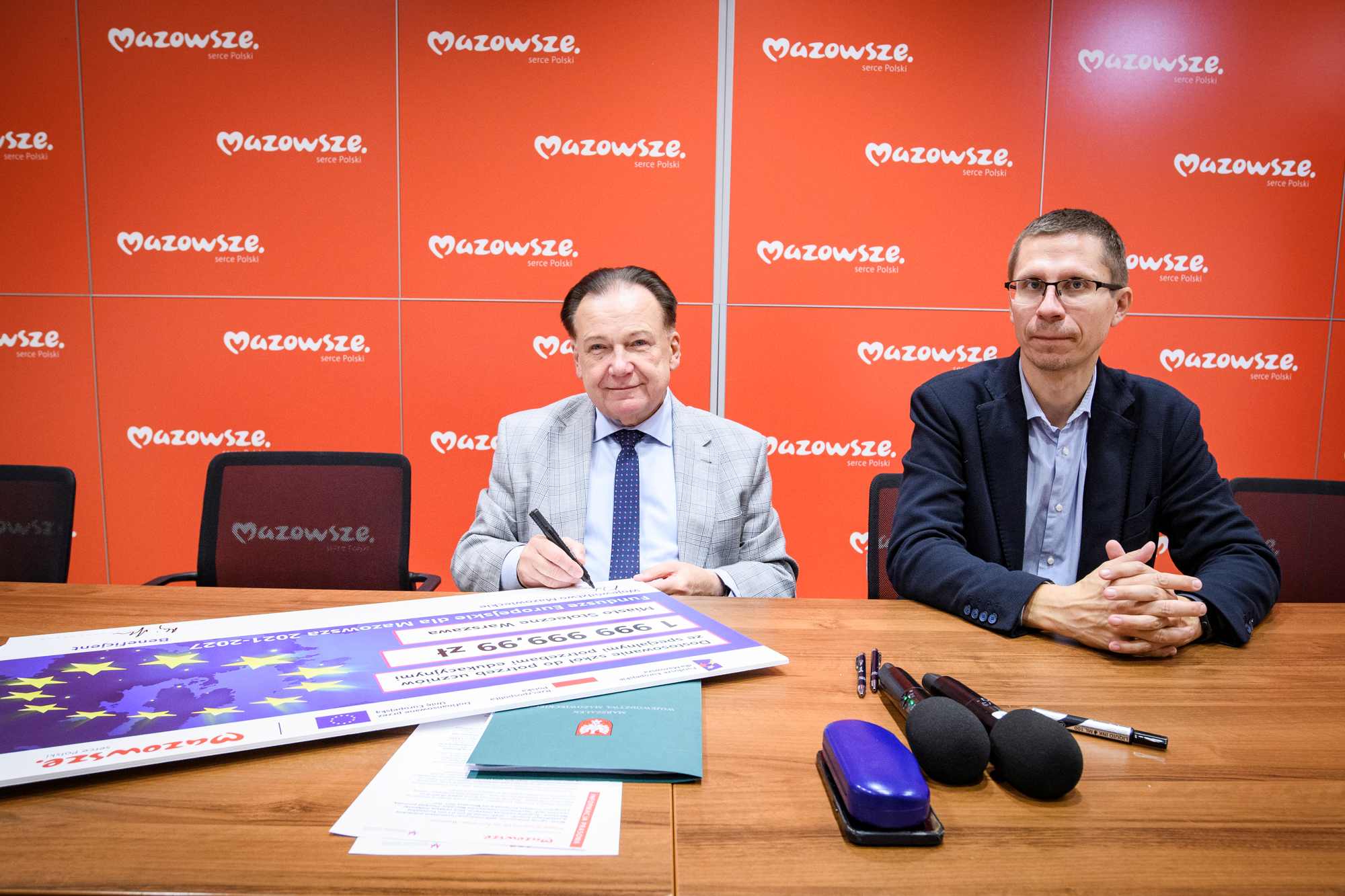 Dwaj mężczyźni siedzą przy drewnianym stole przed pomarańczowym panelem z powtarzającym się białym napisem „Mazowsze serce Polski”, starszy mężczyzna w jasnej kratowanej marynarce trzyma długopis nad dokumentem, obok niego mężczyzna w ciemnej marynarce i okularach trzyma splecione ręce, na stole leży duży plakat z gwiazdami UE, mikrofony, długopisy, etui na okulary i zielona teczka z godłem.