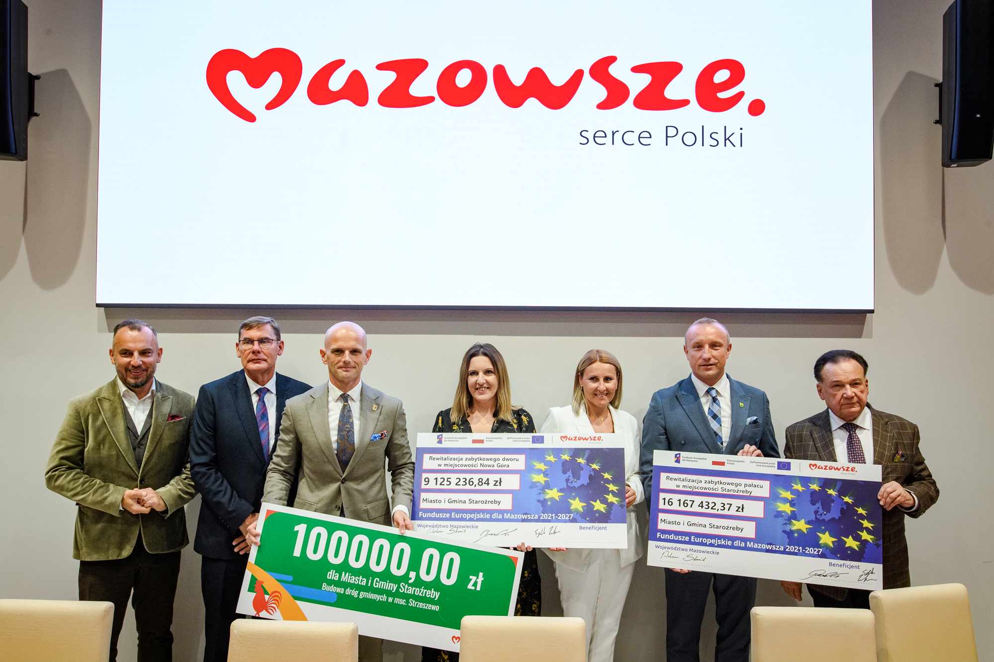 Grupa siedmiu dorosłych stoi w rzędzie przed dużym białym ekranem z czerwonym napisem Mazowsze. serce Polski, trzymając kolorowe, duże czeki symboliczne z motywem gwiazd UE i podanymi kwotami w złotych, w jasnej sali konferencyjnej.