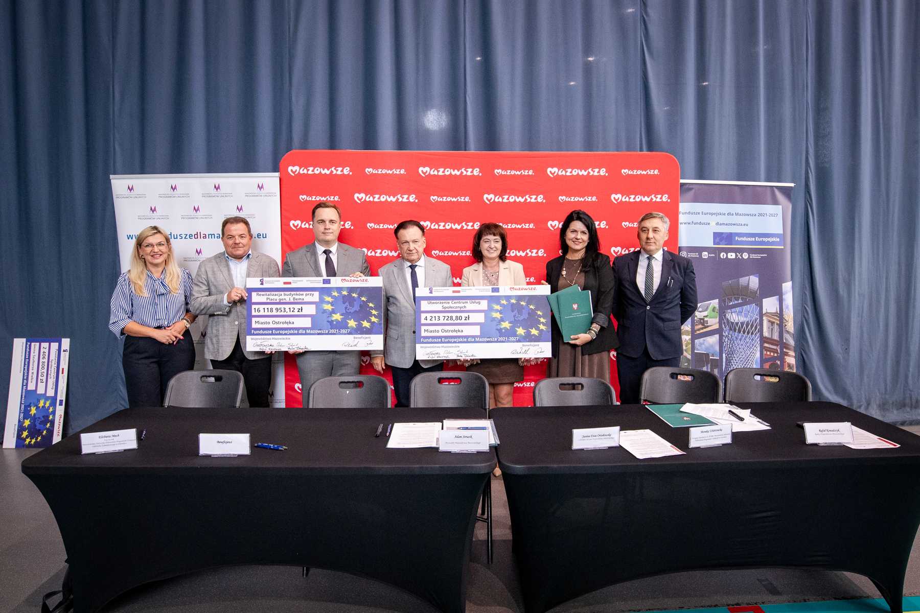 Siedem osób w formalnym ubiorze stoi w rzędzie za czarnym stołem; kilka z nich trzyma dwie duże symboliczne czeki z motywem gwiazd Unii Europejskiej, za grupą widoczny czerwony baner z powtarzającym się napisem "Mazowsze" i niebieskie zasłony, po bokach stoją dodatkowe roll-upy, a na stole leżą dokumenty, długopisy i identyfikatory.