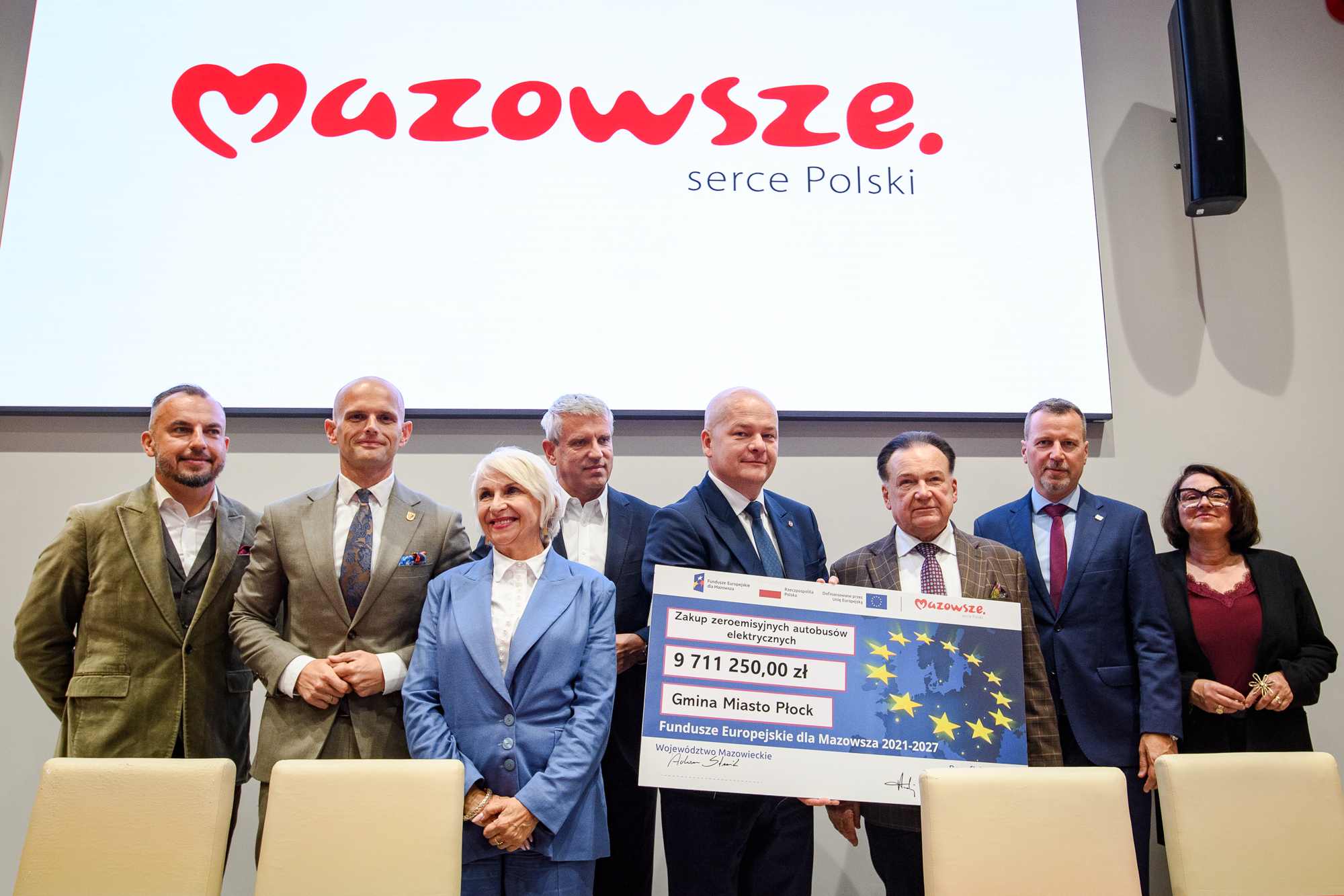 Grupa ośmiu osób w formalnych strojach stoi u dołu kadru przed dużym białym ekranem z czerwonym napisem "Mazowsze serce Polski", trzymając częściowo widoczny duży czek z flagą UE i kwotą 9 711 250,00 zł; na pierwszym planie dwie beżowe fotele, po prawej czarny głośnik na ścianie.