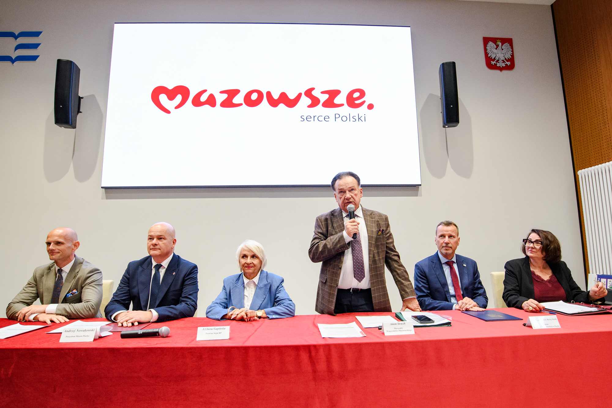 Panel sześciu osób siedzących za stołem z czerwonym obrusem, z mężczyzną stojącym pośrodku trzymającym mikrofon; w tle duży biały ekran z czerwonym napisem "Mazowsze. serce Polski", biało-czerwone godło Polski na ścianie, czarne głośniki po bokach oraz dokumenty i tabliczki z nazwiskami na stole.