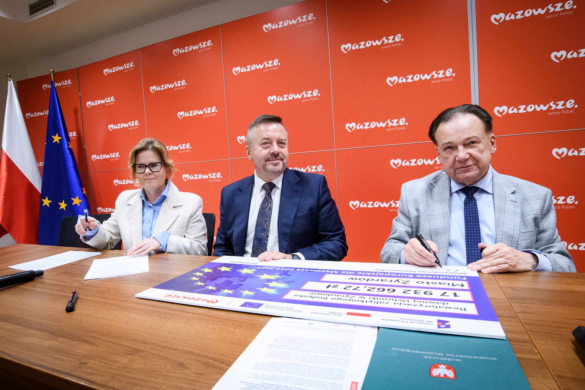 Trzy osoby siedzą przy stole i podpisują dokumenty — kobieta w jasnej marynarce z okularami po lewej, mężczyzna w ciemnym garniturze pośrodku i mężczyzna w jasnej marynarce w kratę po prawej, na stole duży plakat z gwiazdami UE, a w tle czerwone panele z powtarzającym się napisem "Mazowsze" oraz stojące po lewej flagi Polski i Unii Europejskiej.