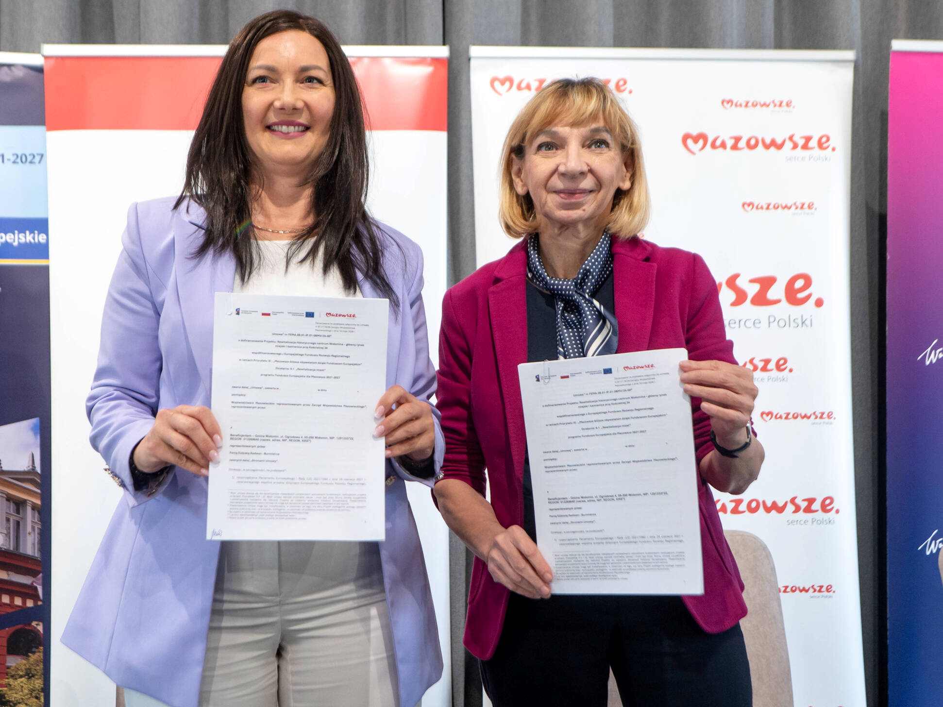 Dwie kobiety stojące obok siebie w połowy kadru, każda trzymająca biały wydrukowany dokument; po lewej w jasnofioletowej marynarce i białej bluzce, po prawej w różowej marynarce i apaszką, w tle pionowe banery z czerwonym napisem "Mazowsze".