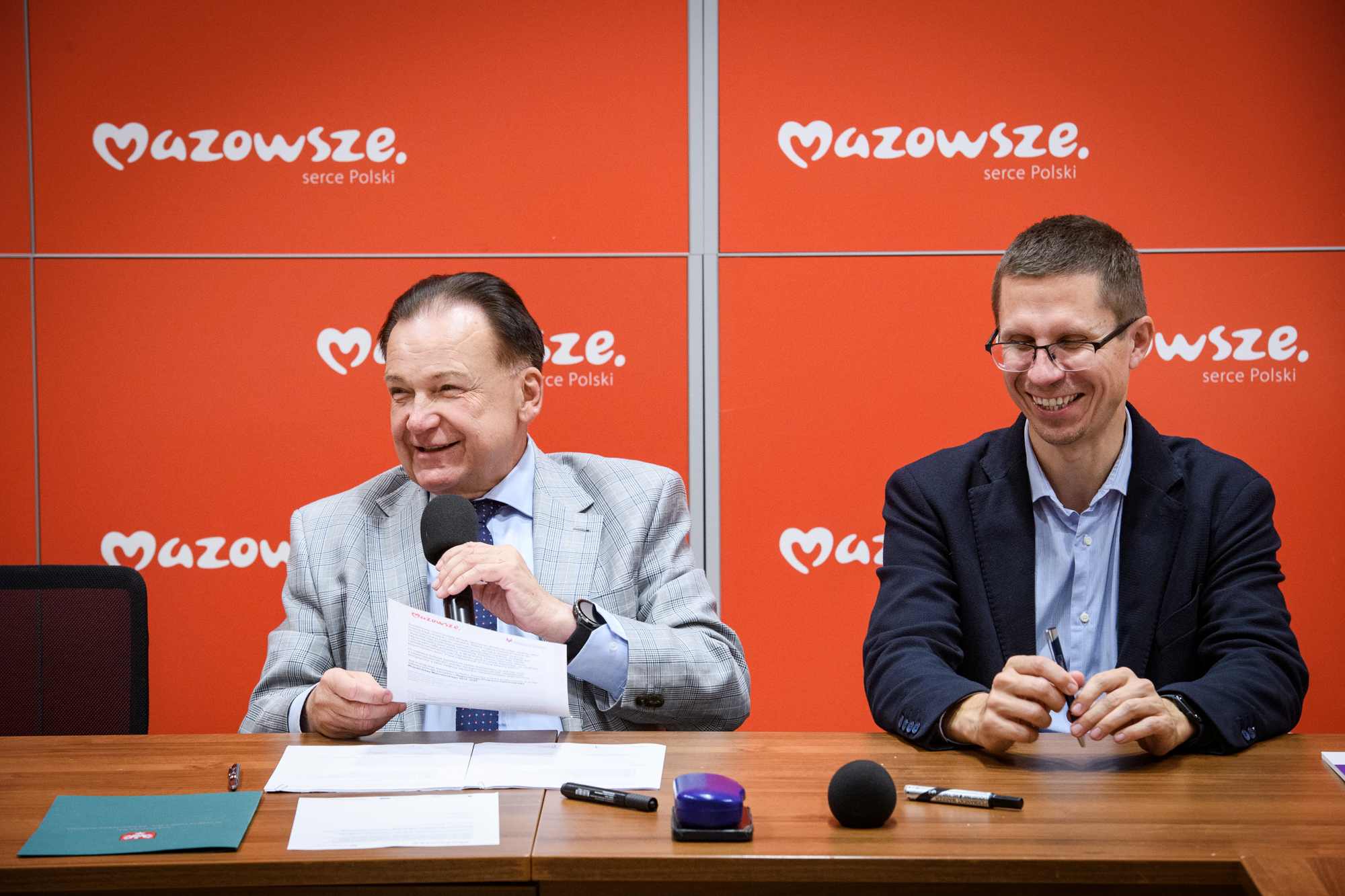 Dwóch mężczyzn siedzi za drewnianym stołem przed czerwonymi panelami z białym logotypem „Mazowsze serce Polski”; po lewej starszy mężczyzna w jasnej marynarce trzyma mikrofon i dokument, po prawej młodszy mężczyzna w okularach i ciemnej marynarce trzyma długopis i się uśmiecha, na stole leżą dokumenty, długopisy, dodatkowy mikrofon i fioletowy stempel.