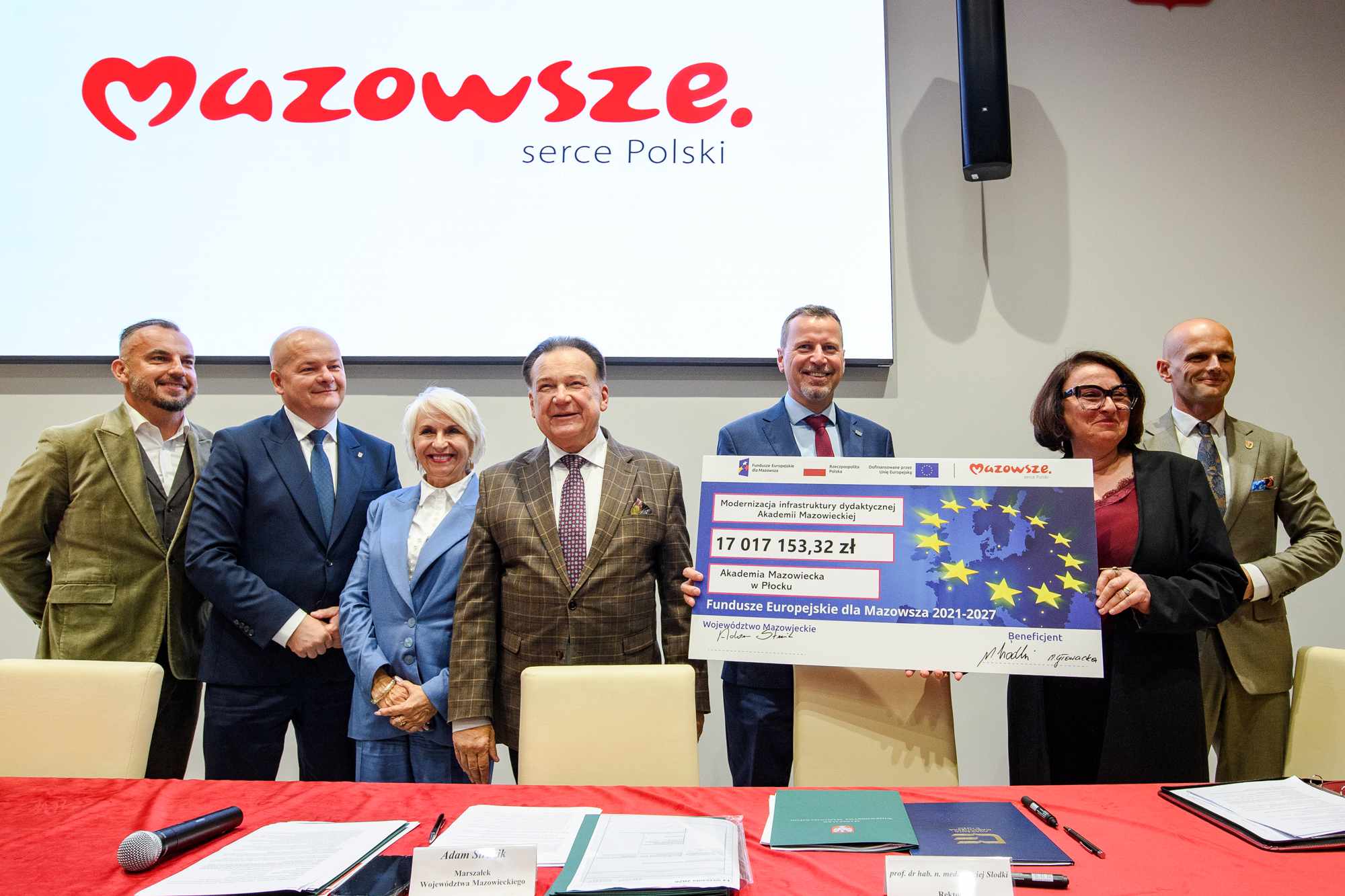 Siedmioro dorosłych w strojach formalnych stoi na scenie przy dużym symboliczny czeku z napisem i gwiazdami Unii Europejskiej oraz kwotą 17 017 153,32 zł, na tle dużego ekranu z czerwonym logo „Mazowsze serce Polski”, a na pierwszym planie czerwony stół z mikrofonem i dokumentami.