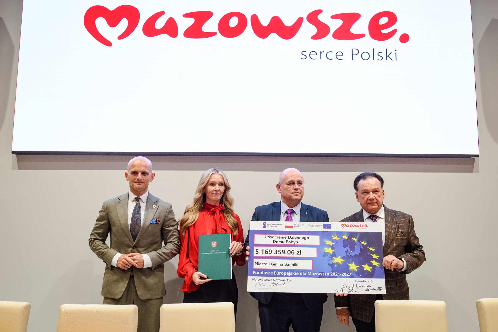 Cztery osoby stoją ramię w ramię pod dużym białym ekranem z czerwonym napisem „Mazowsze serce Polski”: od lewej mężczyzna w jasnoszarym garniturze, kobieta w czerwonej bluzce trzymająca zielony folder, mężczyzna w ciemnym garniturze trzymający duży czek z gwiazdami UE i widoczną kwotą 5 169 359,06 zł oraz starszy mężczyzna w brązowej marynarce w kratę, na pierwszym planie widoczne krzesła.