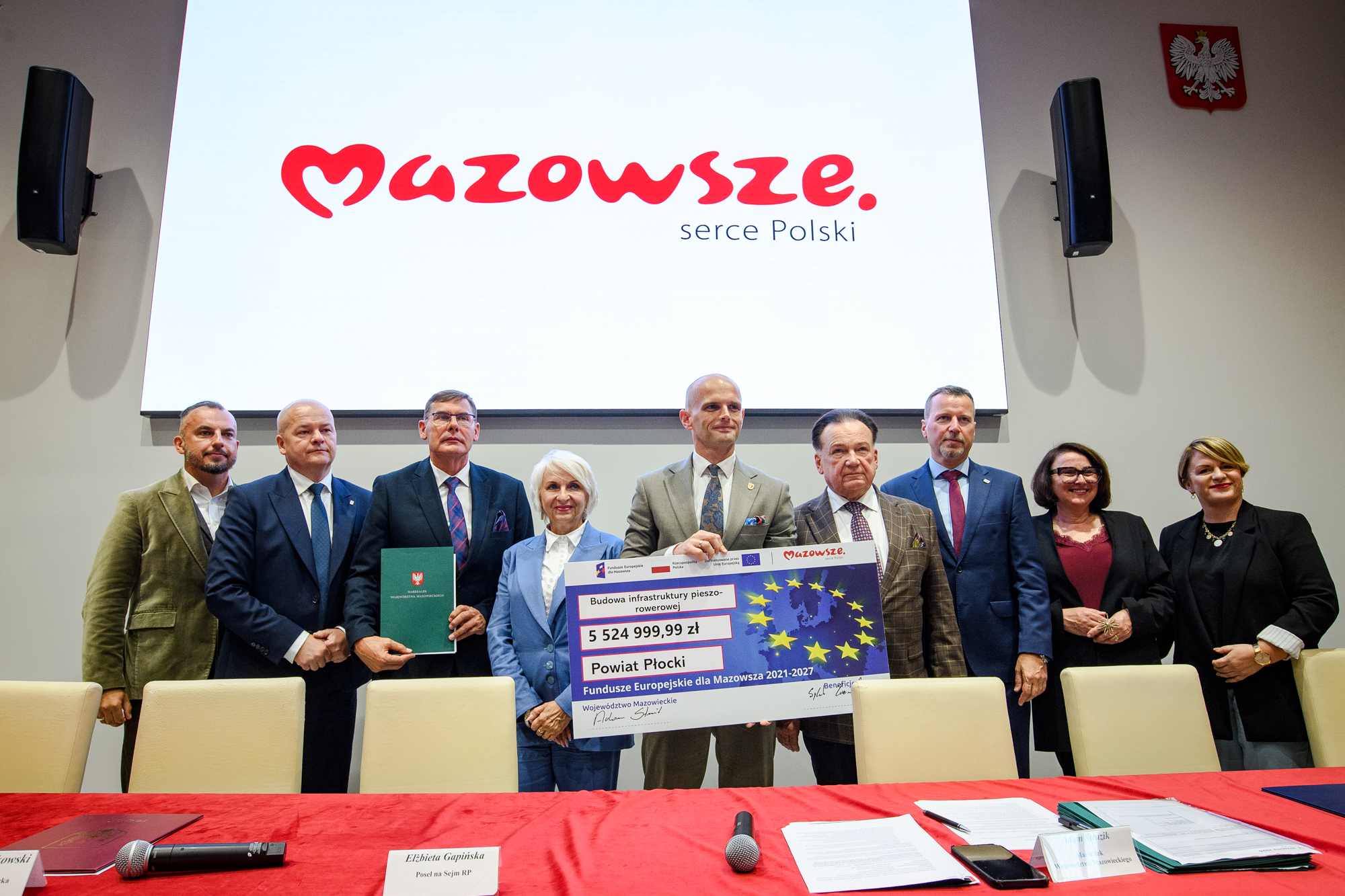 Grupa dziewięciu osób stojących w rzędzie przed dużym białym ekranem z czerwonym logo "Mazowsze" i napisem "serce Polski", kilka osób trzyma duży symboliczny czek z flagą UE, widoczną kwotą 5 524 999,99 zł i napisem "Powiat Płocki", na pierwszym planie stół z czerwonym obrusem, mikrofonami i kartami imiennymi, a na ścianie po prawej widoczne godło Polski.