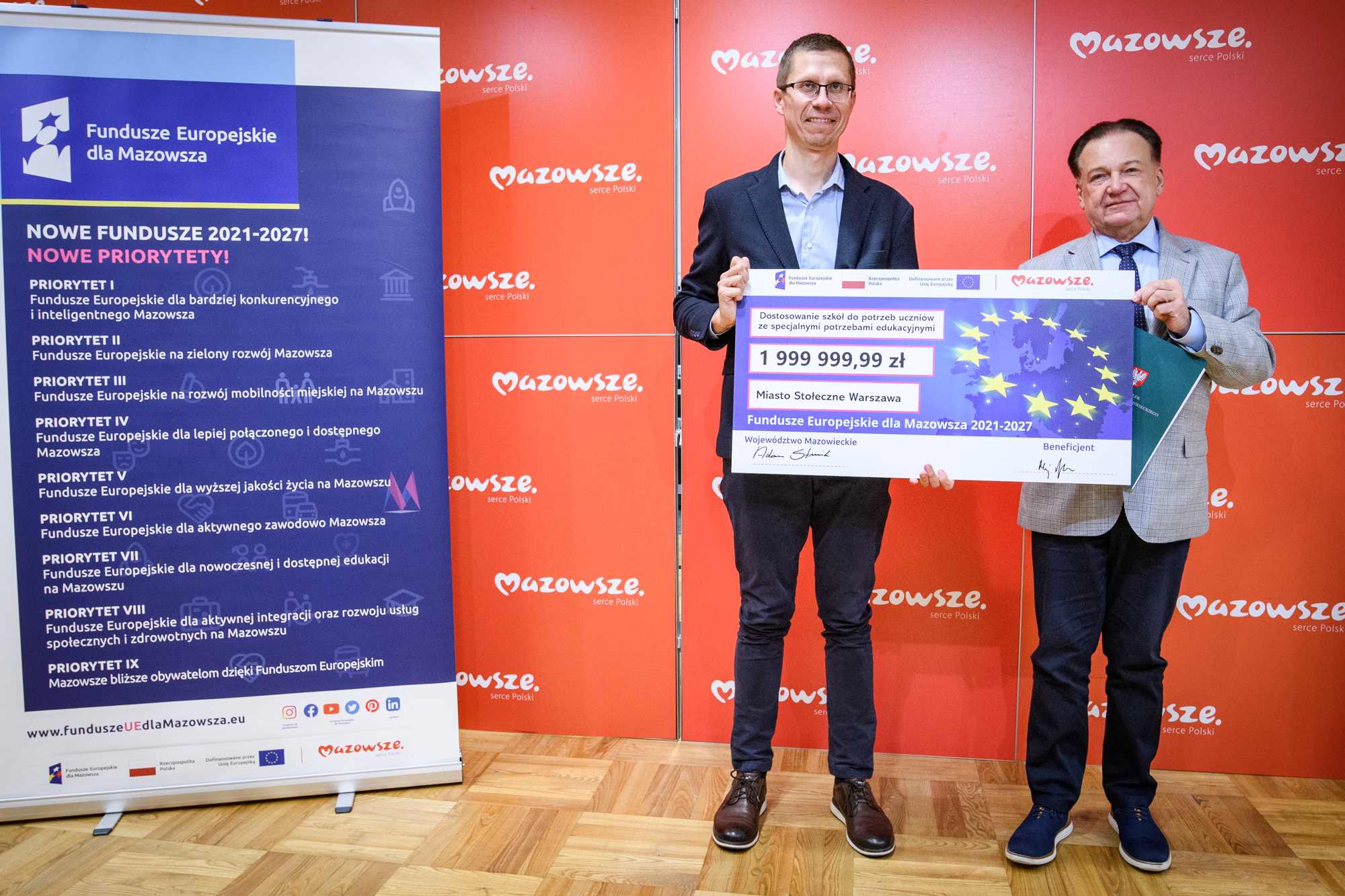 Dwóch mężczyzn stojących na drewnianej podłodze przed czerwonym panelem z napisem „Mazowsze serce Polski”, trzymających duży symboliczny czek na 1 999 999,99 zł z grafiką gwiazd UE i napisem „Miasto Stołeczne Warszawa”, obok niebieski roll‑up „Fundusze Europejskie dla Mazowsza” z listą priorytetów.
