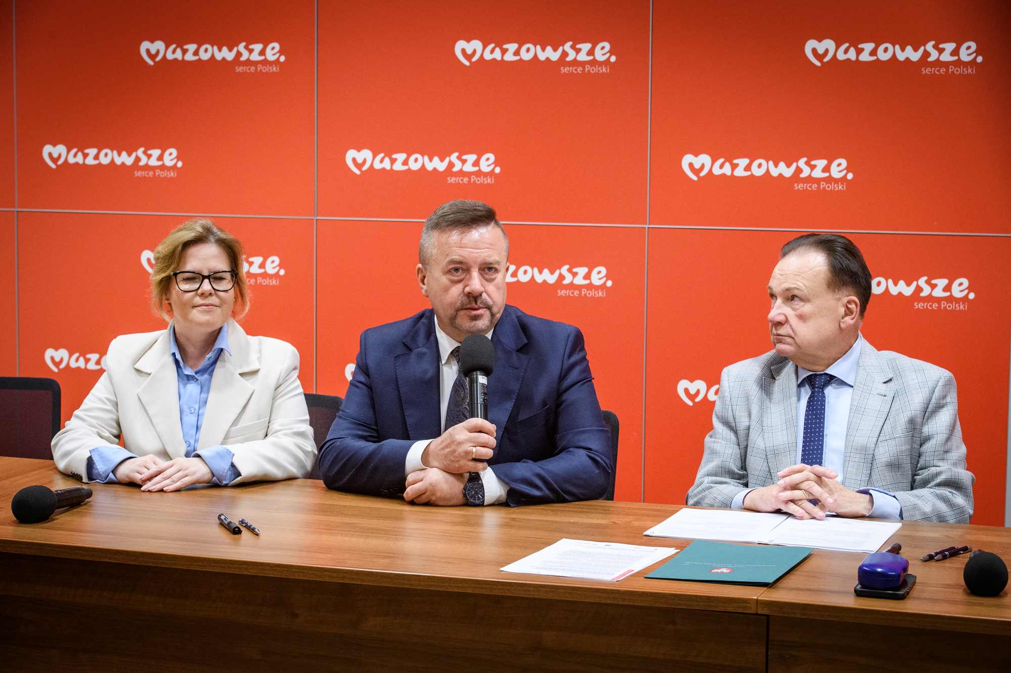 Trzy osoby siedzą przy drewnianym stole na tle czerwonych paneli z białym napisem „Mazowsze serce Polski”: środkowy mężczyzna w ciemnym garniturze trzyma mikrofon, po lewej kobieta w jasnej marynarce i okularach, po prawej mężczyzna w jasnym kratkowanym garniturze, na stole dokumenty, długopisy i dodatkowy mikrofon.