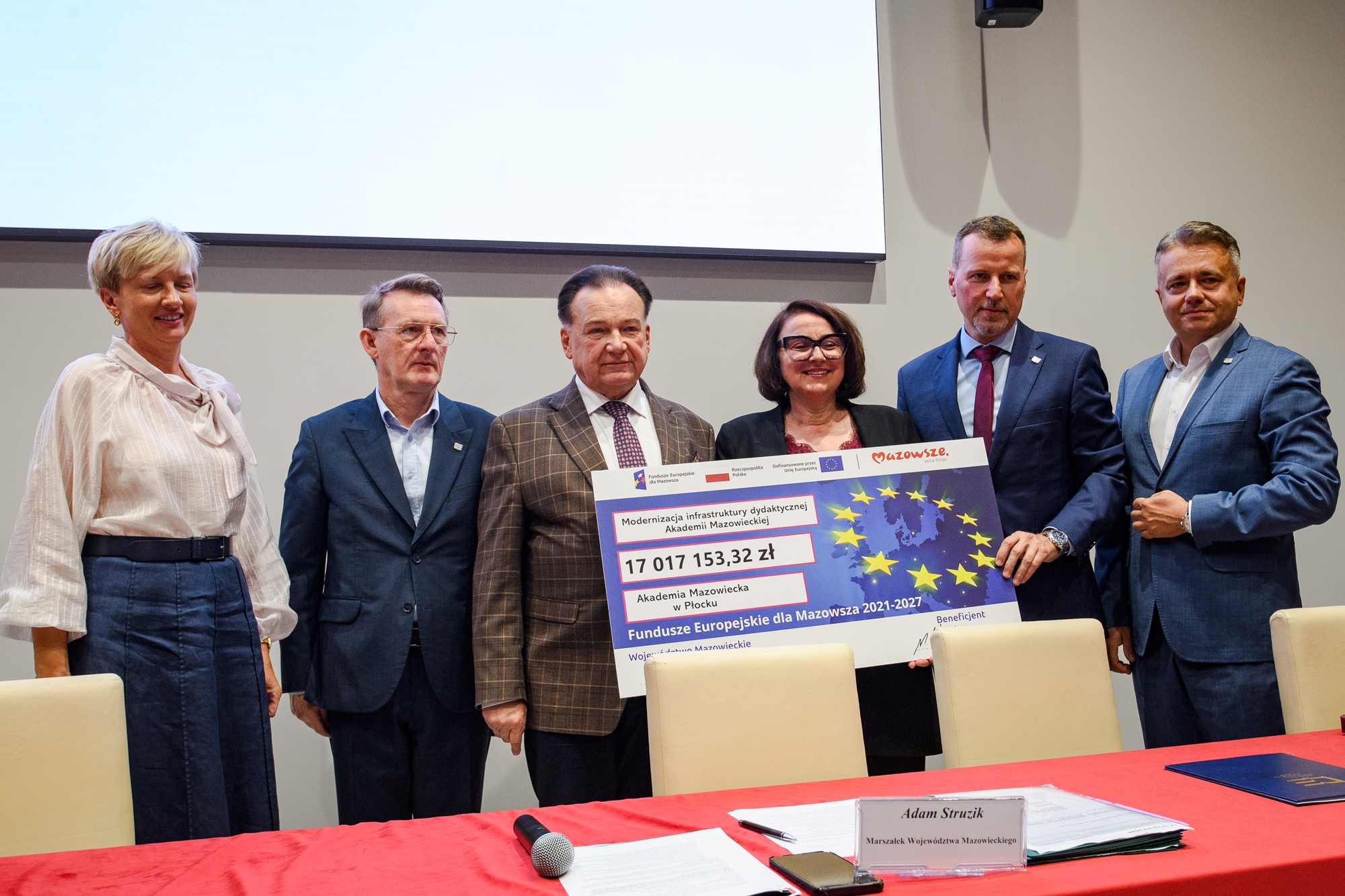 Sześć osób w formalnym ubiorze stoi w rzędzie przed dużym białym ekranem trzymając wielki symboliczny czek z motywem gwiazd UE i widoczną kwotą 17 017 153,32 zł, a na pierwszym planie czerwony stół z mikrofonem, dokumentami i plastikową tabliczką z napisem.