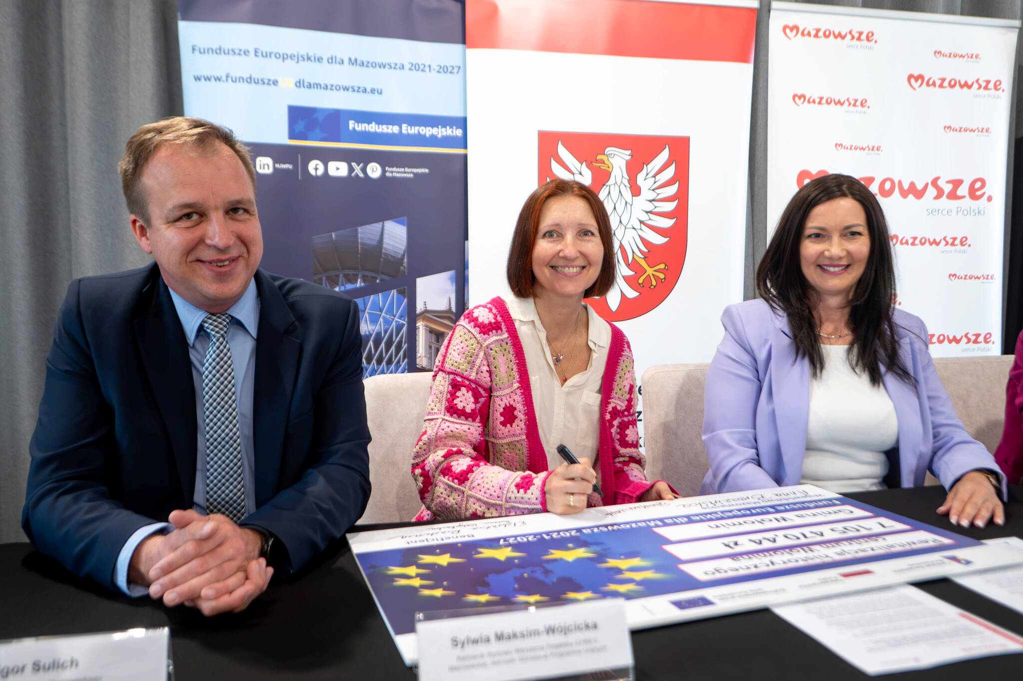 Troje osób siedzi przy stole podpisując dokumenty: po lewej mężczyzna w granatowym garniturze i kraciastej krawacie, pośrodku kobieta z rudymi włosami w różowo‑białym szydełkowanym swetrze trzymająca długopis, po prawej kobieta z ciemnymi włosami w jasnofioletowej marynarce; na stole widoczna plansza z flagą UE i dokumenty, w tle banery z białym orłem w czerwonej tarczy i napisem "Mazowsze".