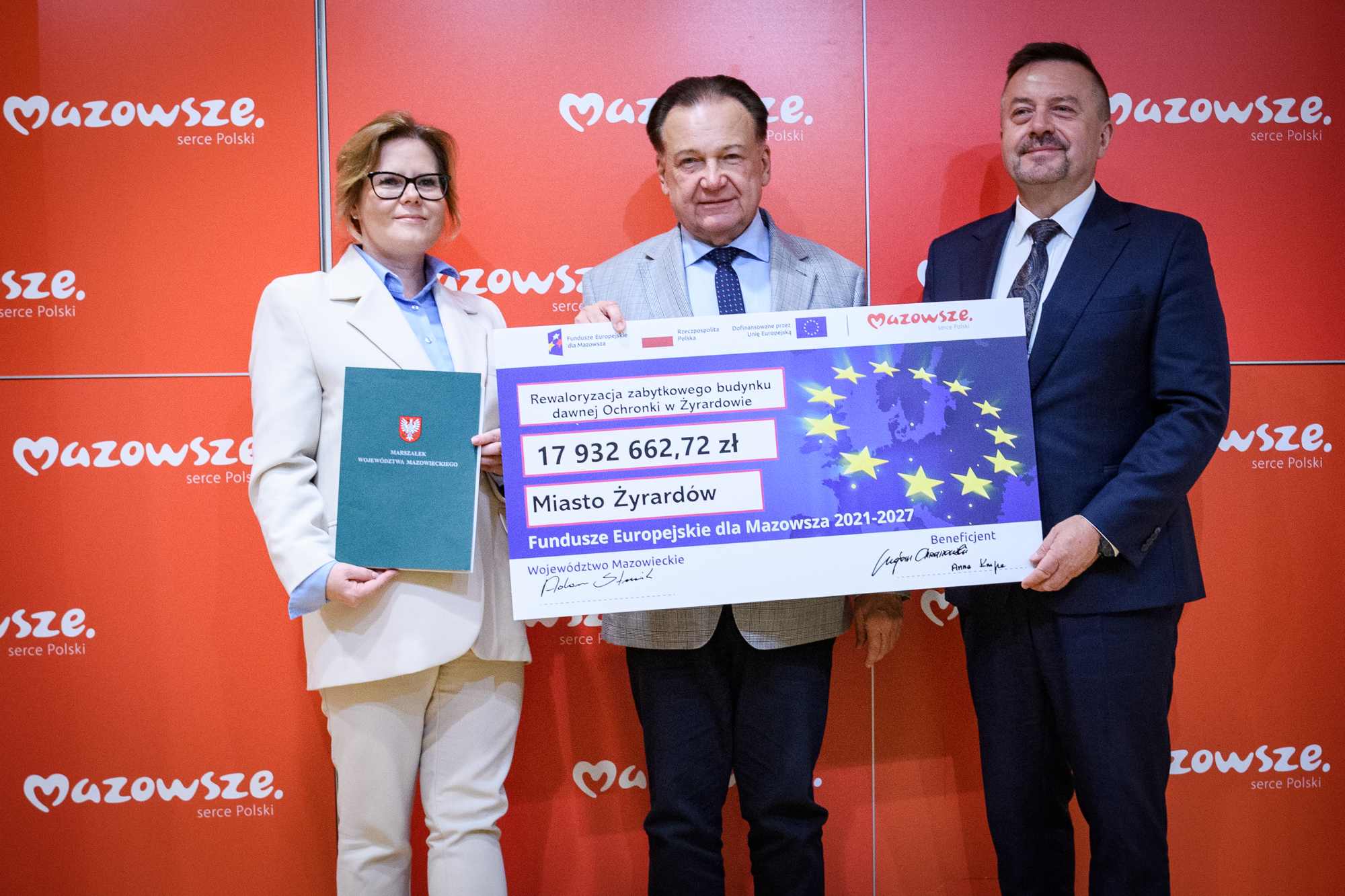 Trzy osoby w formalnych strojach stoją przed czerwonym panelem z powtarzającym się białym napisem „Mazowsze serce Polski” i trzymają duży symboliczny czek z motywem gwiazd UE i kwotą 17 932 662,72 zł; po lewej kobieta w jasnym garniturze i okularach trzyma zielony folder z herbem.
