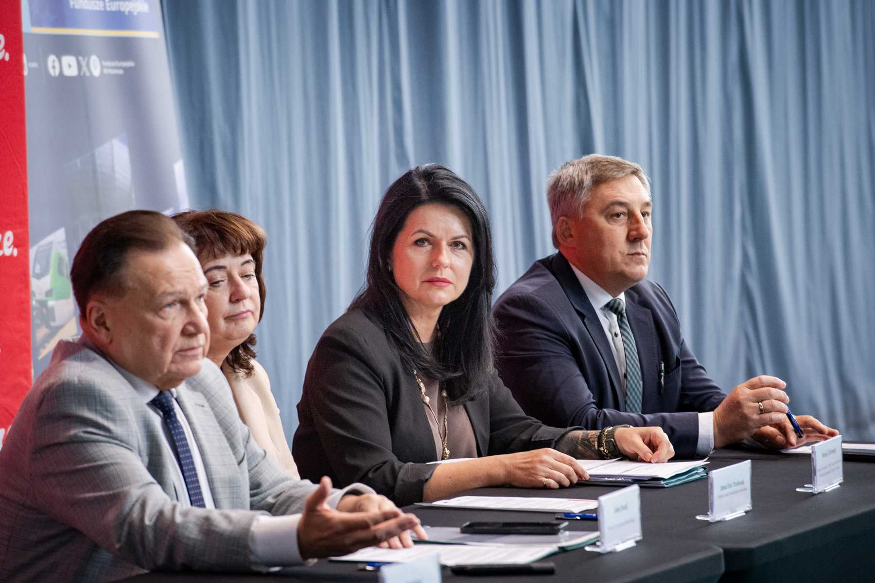 Czterech dorosłych siedzi obok siebie za stołem konferencyjnym — od lewej starszy mężczyzna w jasnej marynarce, kobieta z krótkimi brązowymi włosami, kobieta z długimi czarnymi włosami i mężczyzna w ciemnym garniturze; na stole leżą dokumenty, długopisy i plastikowe tabliczki z nazwiskami, w tle niebieska zasłona i czerwony baner.