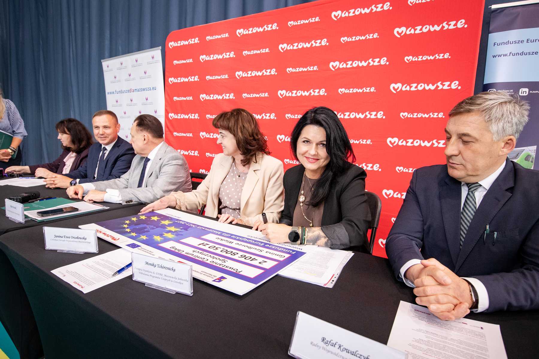 Kilka osób w formalnych strojach siedzi przy długim czarnym stole z dokumentami i dużym planszowym czekiem z motywem flagi UE, jedna kobieta z czarnymi włosami trzyma długopis i uśmiecha się do aparatu, a za nimi czerwone tło z powtarzającym się białym napisem "Mazowsze" oraz roll-upy informacyjne po bokach.