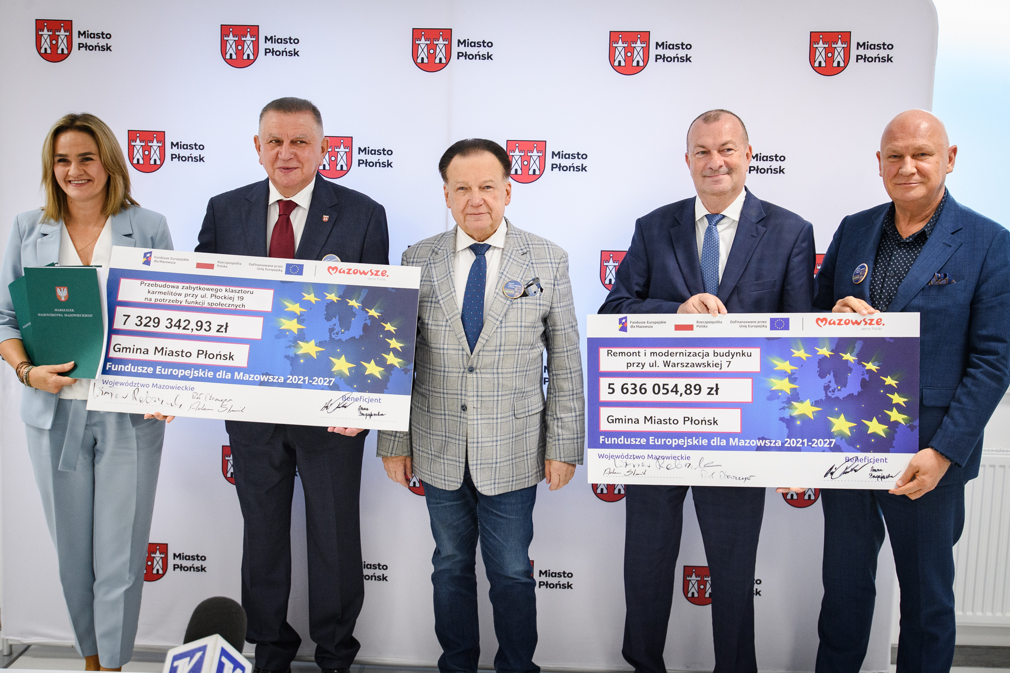 Pięć osób w formalnych strojach stoi w rzędzie przed białym panelem z herbem i napisem "Miasto Płońsk", dwie osoby trzymają duże czeki z motywem gwiazd UE i widocznymi kwotami 7 329 342,93 zł i 5 636 054,89 zł, kobieta po lewej trzyma zielony akt, a u dołu kadru widoczny mikrofon telewizyjny.