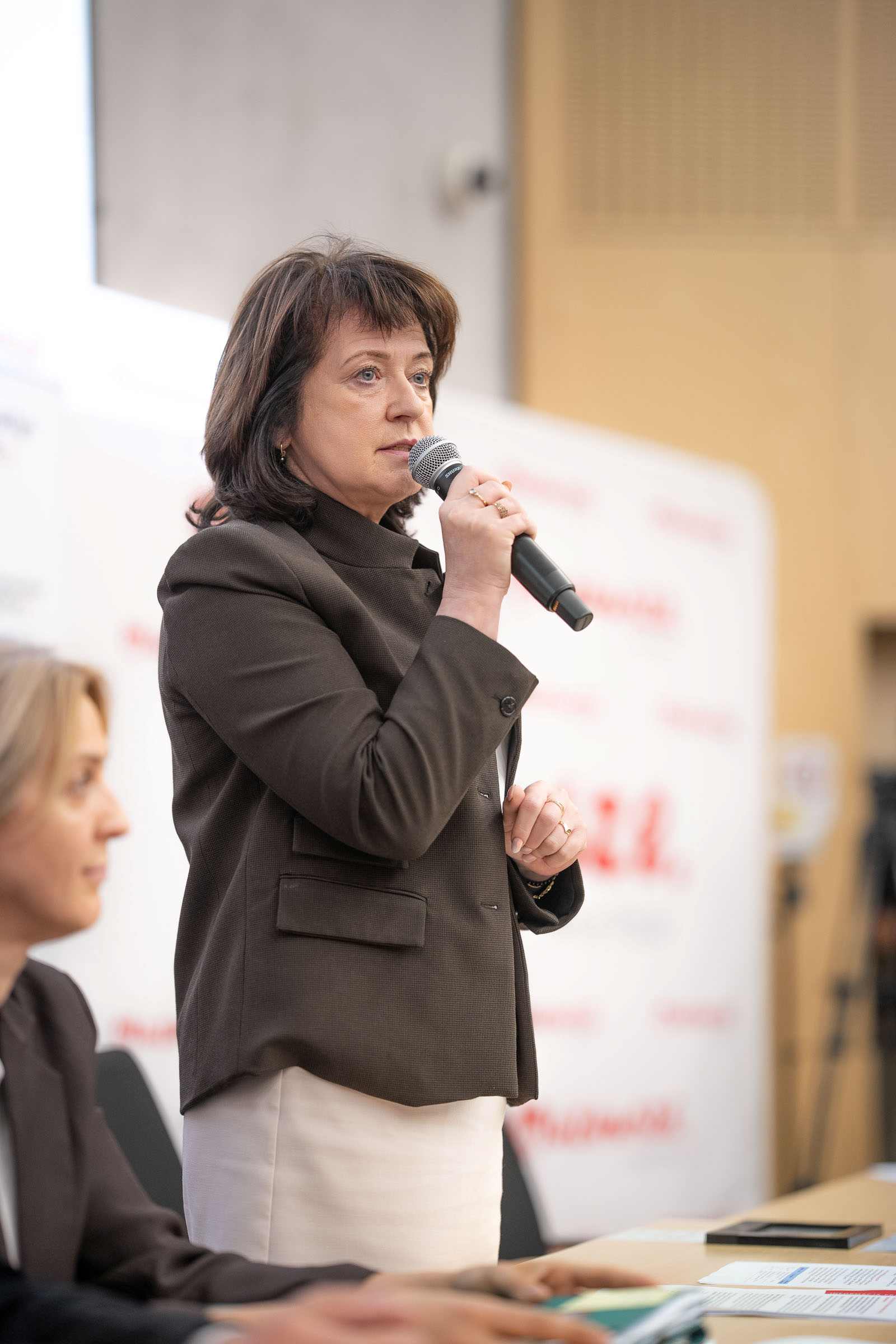 Kobieta w średnim wieku z brązowymi włosami trzyma mikrofon przy ustach, stoi na sali konferencyjnej w ciemnej marynarce i jasnej spódnicy; z przodu rozmyta sylwetka innej osoby, w tle rozmyty biały baner z czerwonym napisem i stół z dokumentami.