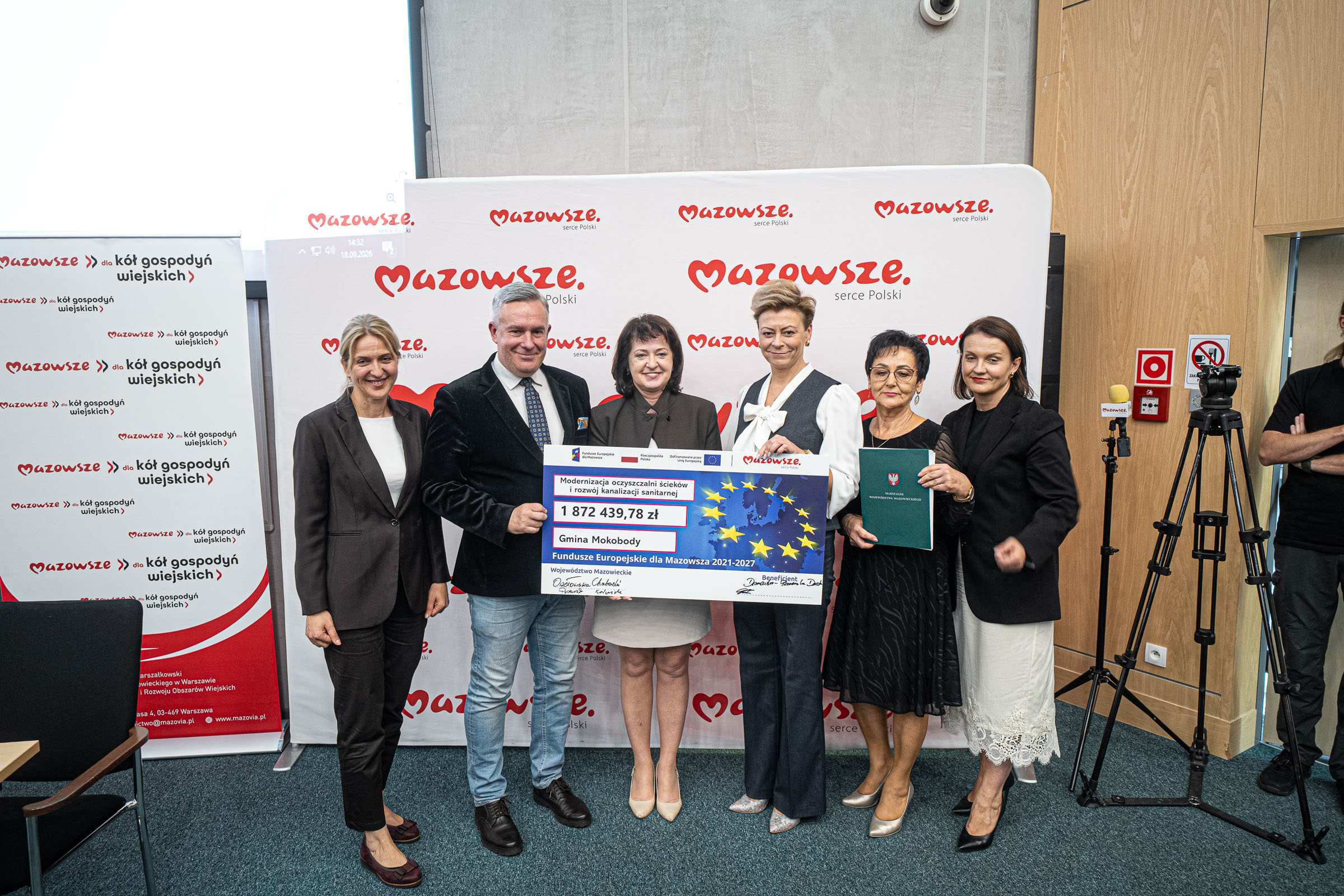 Grupowe zdjęcie sześciu dorosłych stojących na wykładzinie przed białym banerem z czerwonym logo „Mazowsze”, trzy osoby trzymają duży symboliczny czek z napisem „1 872 439,78 zł” i „Gmina Mokobody”, po lewej widoczny pionowy baner z napisem „dla kół gospodyń wiejskich”, a po prawej kamera na statywie i drewniana ściana.