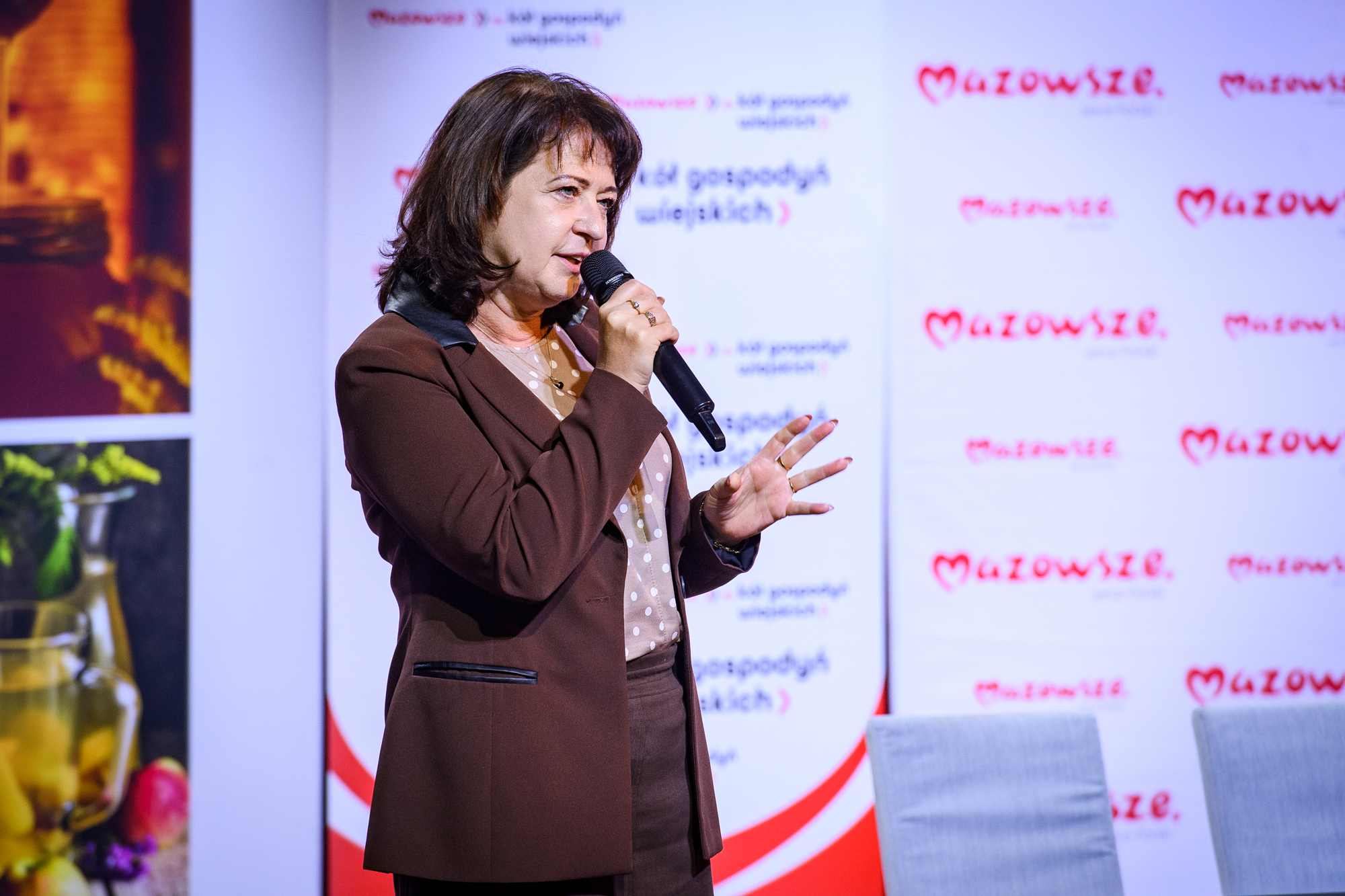 Kobieta w brązowym garniturze i bluzce w kropki trzyma mikrofon i gestykuluje na tle białych banerów z czerwonym napisem „Mazowsze” oraz szarych foteli, po lewej rozmyte zdjęcia słoików i owoców.