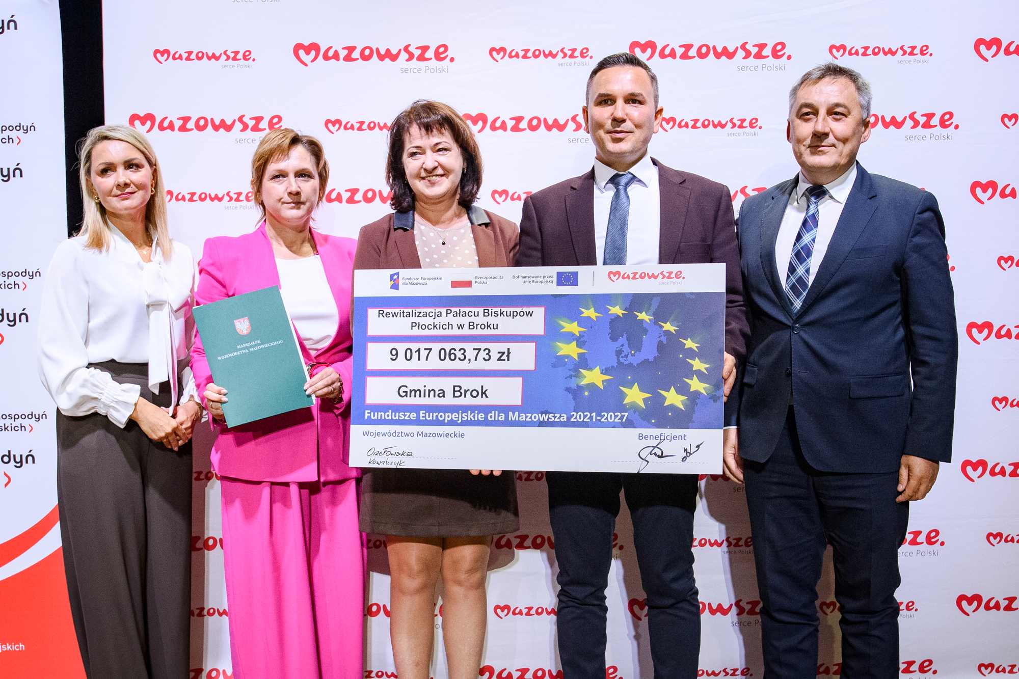 Pięć osób stojących obok siebie przed białym panelem z powtarzającym się czerwonym logo "Mazowsze", trzymających duży symboliczny czek z motywem gwiazd Unii Europejskiej i napisem „Rewitalizacja Pałacu Biskupów Płockich w Broku” oraz kwotą 9 017 063,73 zł; od lewej kobieta w białej bluzce i ciemnych spodniach, kobieta w różowym garniturze trzymająca zielony folder, kobieta w brązowej marynarce, mężczyzna w ciemnym garniturze z krawatem i mężczyzna w granatowym garniturze z kraciastą krawatą.