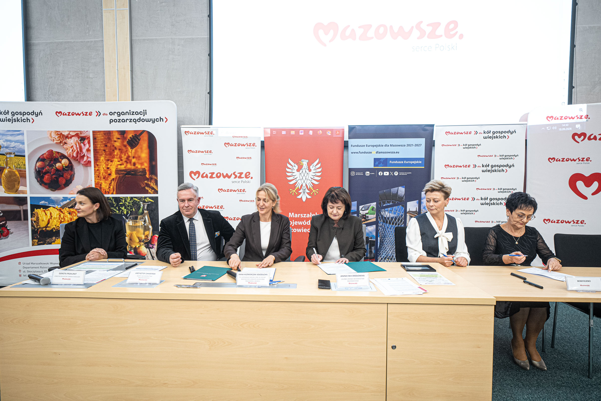 Sześć osób w formalnych strojach siedzi przy długim, jasnym stole i podpisuje dokumenty, przed nimi leżą teczki, długopisy i tabliczki z nazwiskami, a w tle ustawione są pionowe banery promocyjne z powtarzającym się czerwonym napisem "Mazowsze", herbem państwowym z orłem oraz zdjęciami żywności i informacji o funduszach.