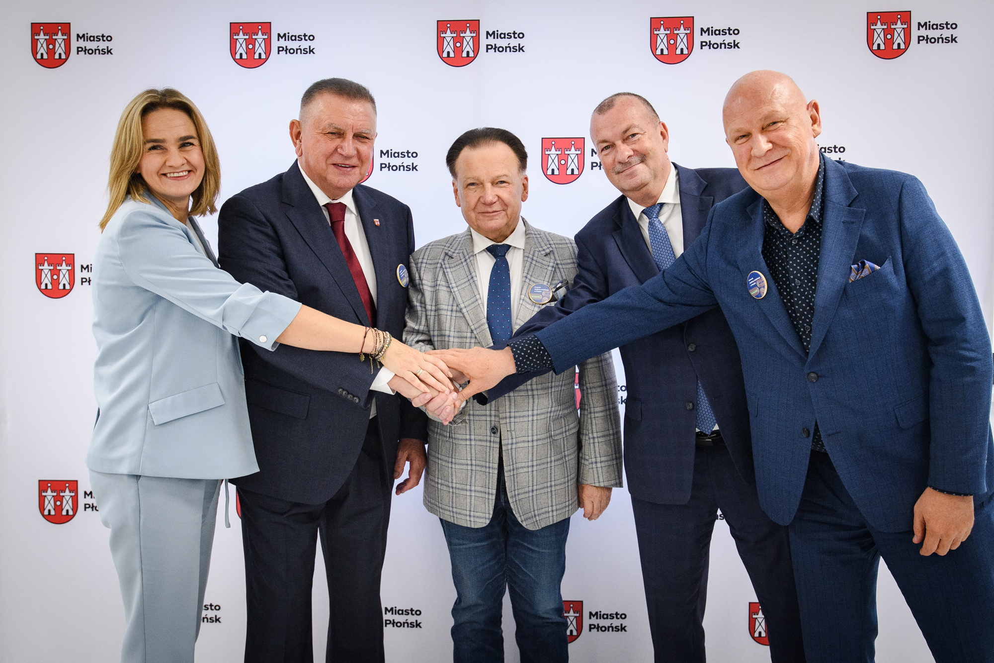 Pięć osób w oficjalnych marynarkach stoi przed białym tłem z powtarzającym się czerwonym herbem i napisem „Miasto Płońsk”, uśmiechnięci łączą dłonie na środku — kobieta po lewej w jasnobłękitnym garniturze, trzej mężczyźni w ciemnych garniturach i mężczyzna w szarej kratowanej marynarce.