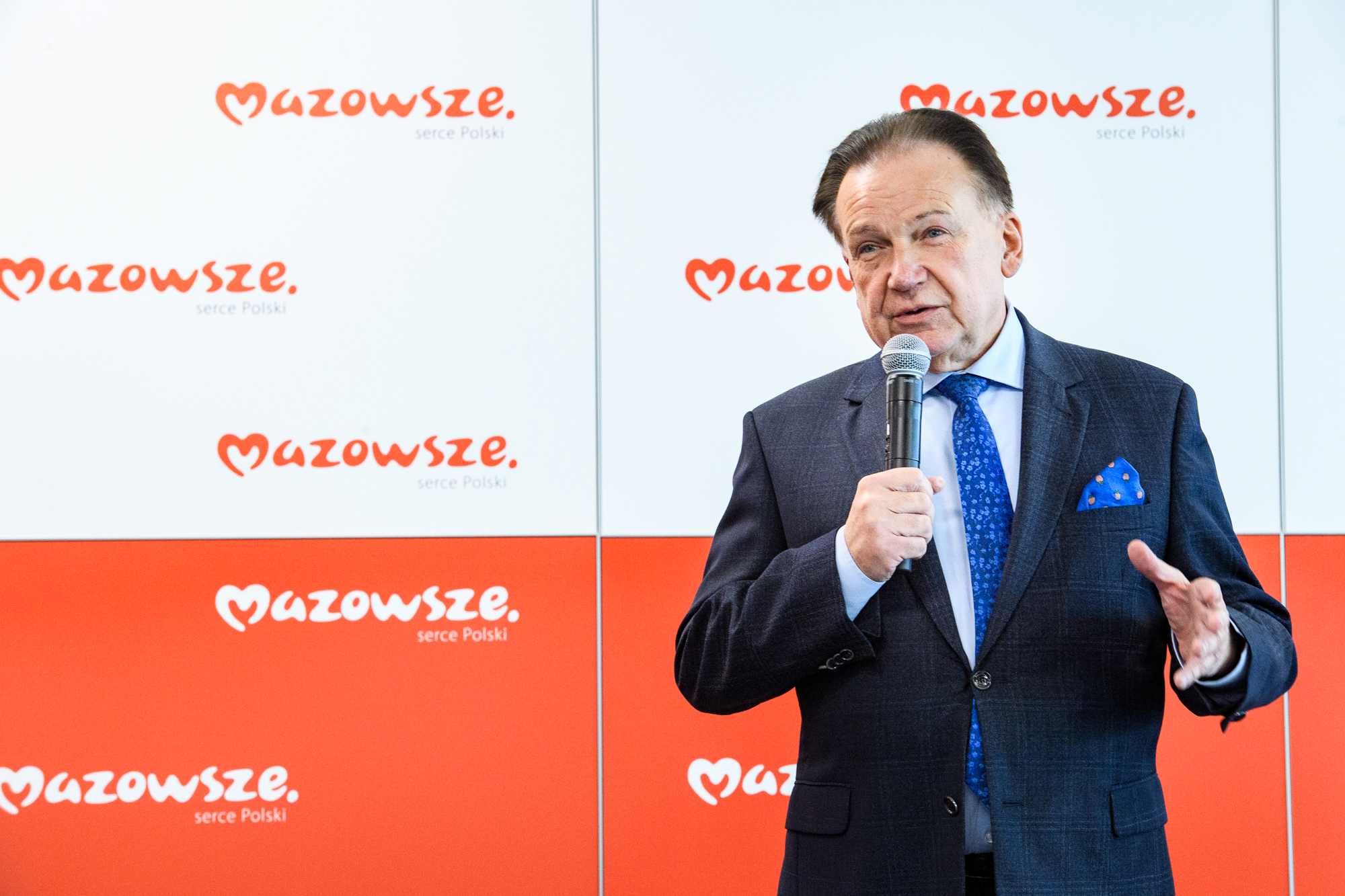 Po prawej stronie kadru mężczyzna w granatowym garniturze z niebieskim krawatem i poszetką trzyma mikrofon i mówi na tle biało‑czerwonych paneli z powtarzającym się logo „Mazowsze. serce Polski”.