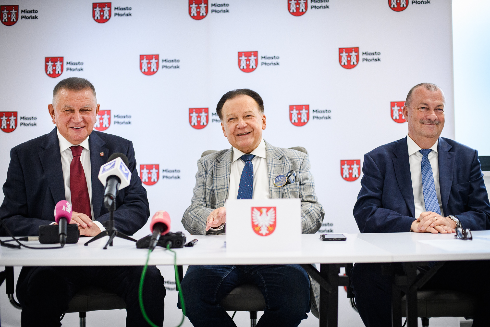 Trzech mężczyzn w garniturach siedzi za stołem z mikrofonami, na środku mężczyzna w szarej kratowanej marynarce z przypiętym znaczkiem, po bokach mężczyźni w ciemnych garniturach z bordowym i niebieskim krawatem, na stole kartka z czerwono-białym herbem, a w tle powtarzający się biały baner z czerwonym herbem i napisem "Miasto Płońsk".