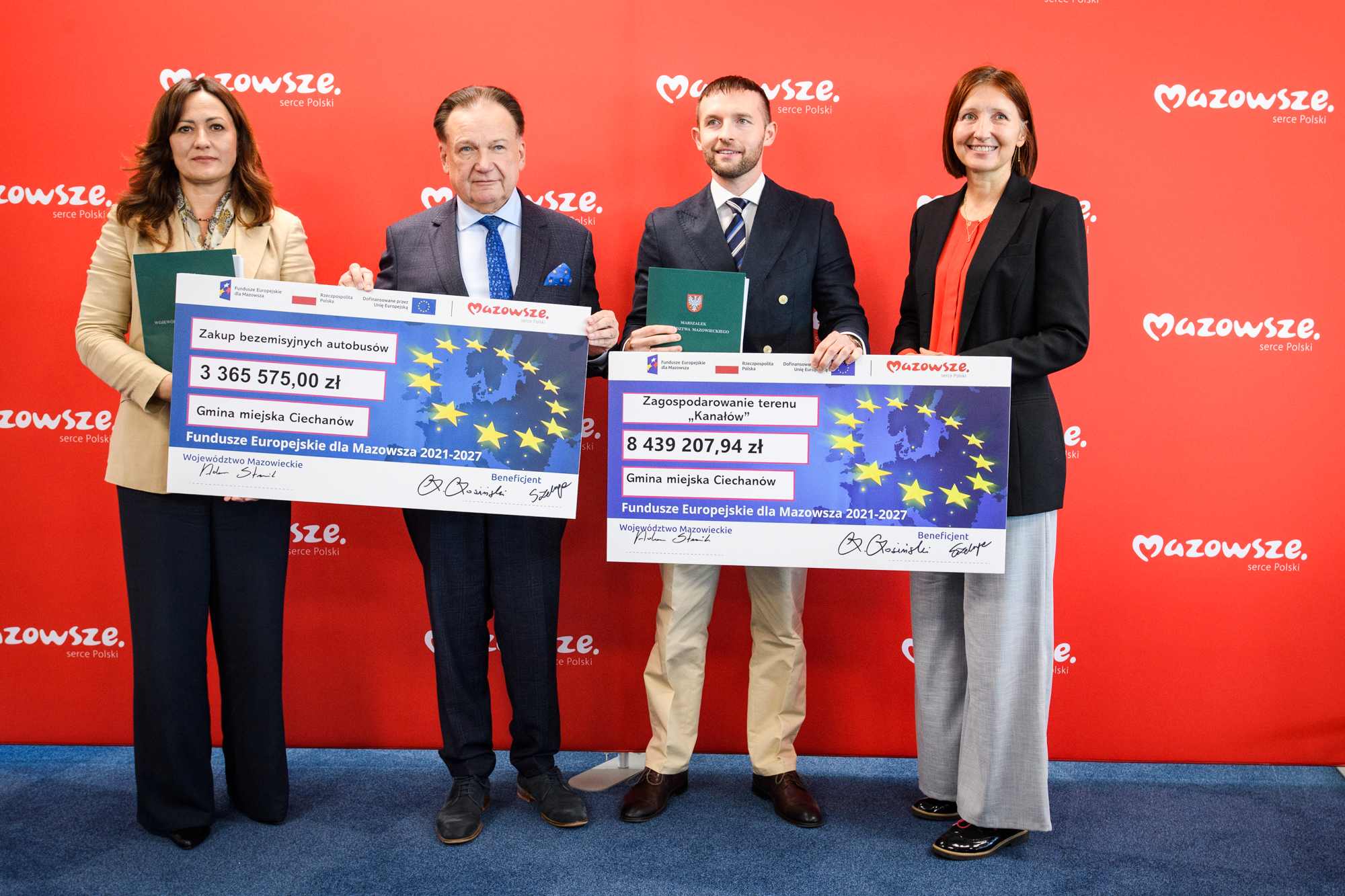 Na czerwonym tle z powtarzającym się logo "Mazowsze serce Polski" stoją w rzędzie cztery osoby — od lewej kobieta w beżowej marynarce trzymająca zielony folder i duży symboliczny czek z napisem "Zakup bezemisyjnych autobusów" na kwotę 3 365 575,00 zł, dalej mężczyzna w ciemnym garniturze z niebieskim krawatem, obok niego mężczyzna w ciemnej marynarce i krawacie trzymający drugi czek z napisem "Zagospodarowanie terenu 'Kanałów'" na kwotę 8 439 207,94 zł oraz z prawej kobieta w czarnej marynarce i czerwonej b