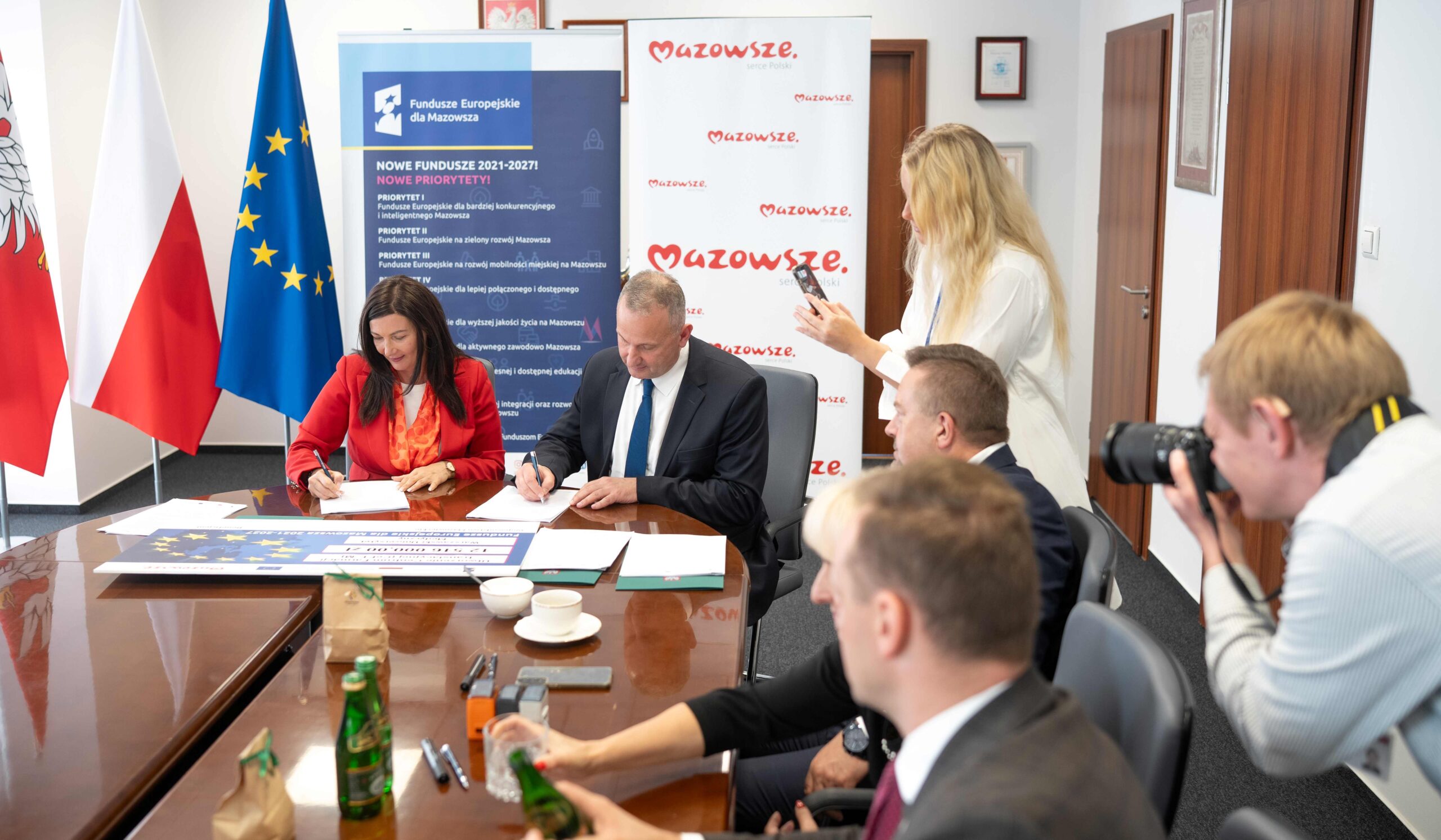 Kilka osób siedzących przy błyszczącym stole konferencyjnym podpisuje dokumenty; po lewej stoją flagi Polski i Unii Europejskiej oraz roll-upy z napisami Mazowsze i Fundusze Europejskie dla Mazowsza, po prawej kobieta robi zdjęcie telefonem, z przodu fotograf używa aparatu, a na stole leżą butelki, filiżanki, długopisy i duży plakat z gwiazdami UE.
