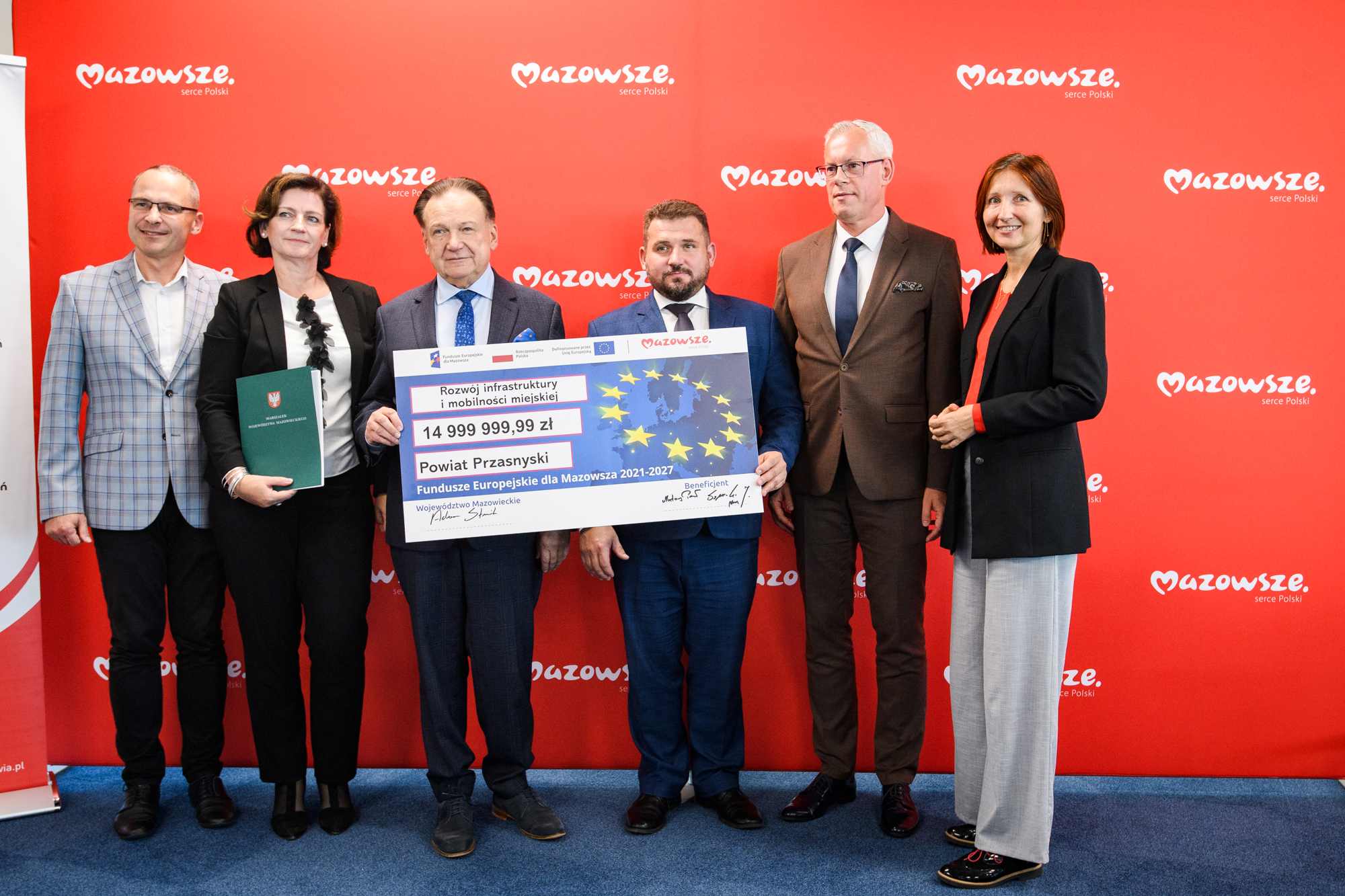 Sześć osób w ubraniach formalnych stoi w rzędzie przed czerwonym banerem z powtarzającym się logo „Mazowsze”, kilka z nich trzyma centralnie duży symboliczny czek z motywem gwiazd UE i kwotą 14 999 999,99 zł na rzecz Powiatu Przasnyski, jedna osoba trzyma zieloną teczkę, a pod nogami widoczny jest niebieski dywan.