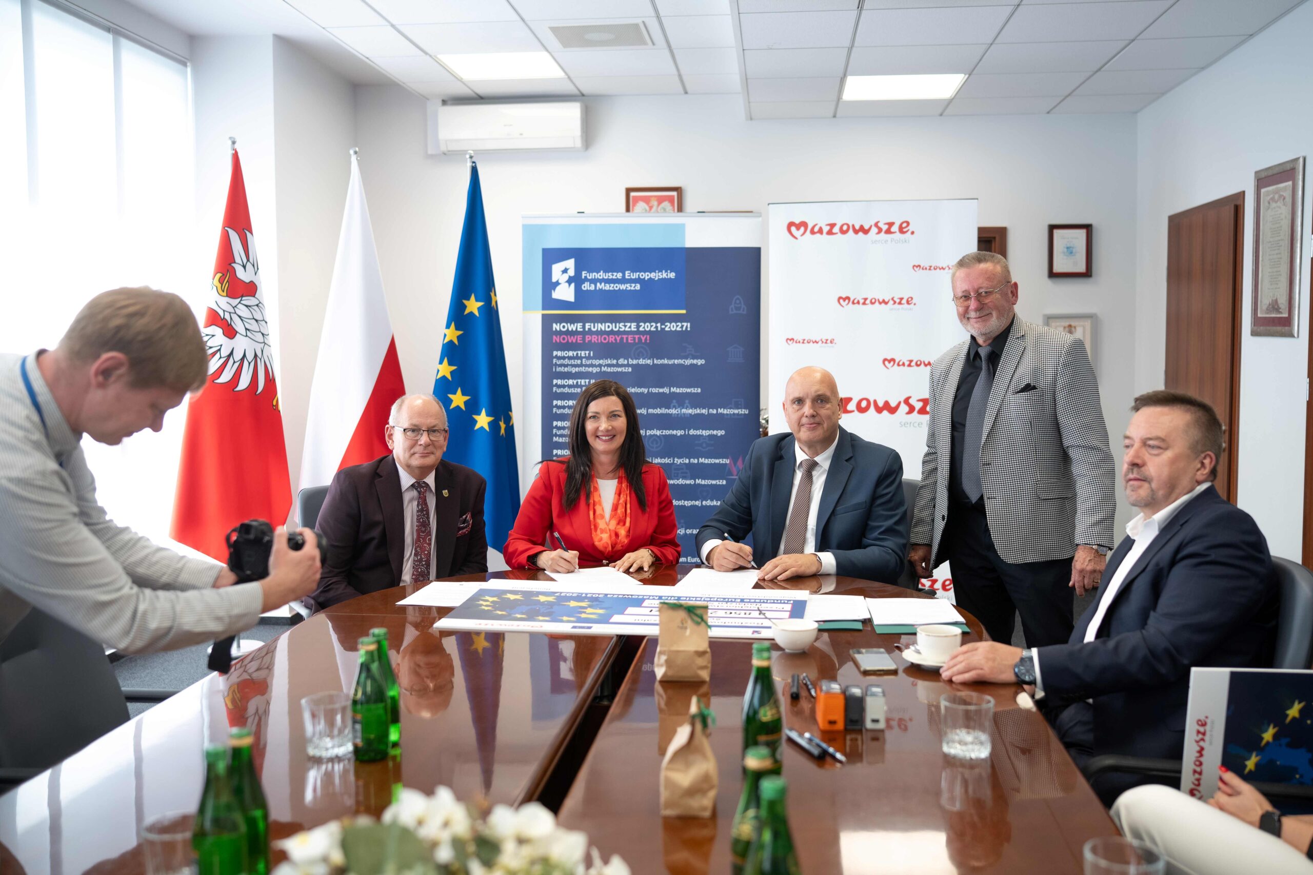 Pokój konferencyjny z grupą osób przy błyszczącym stole: po lewej fotograf z aparatem, przy stole kobieta w czerwonej marynarce i mężczyzna w garniturze podpisują dokumenty, obok siedzą i stoją inni mężczyźni w formalnych ubraniach, w tle flagi Polski i Unii Europejskiej oraz roll-upy z napisami "Fundusze Europejskie dla Mazowsza" i "Mazowsze", na stole dokumenty, butelki wody, filiżanki i bukiet kwiatów.
