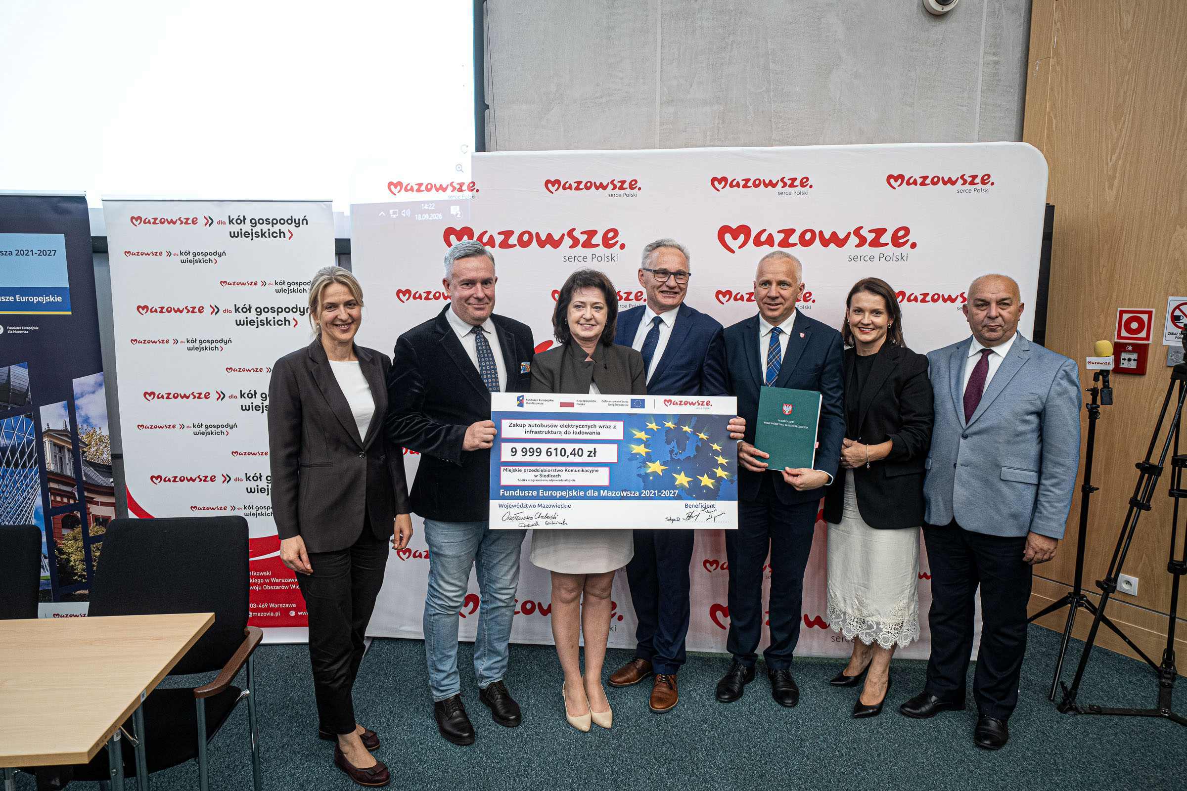 Grupa siedmiu osób w biznesowych strojach stoi w rzędzie przed białym banerem z czerwonym logo „Mazowsze serce Polski”, trzymając duży symboliczny czek z flagą UE i widoczną kwotą 9 999 610,40 zł; po bokach widać dodatkowe roll-upy, statyw z kamerą oraz panel ścienny z przyciskiem alarmowym.