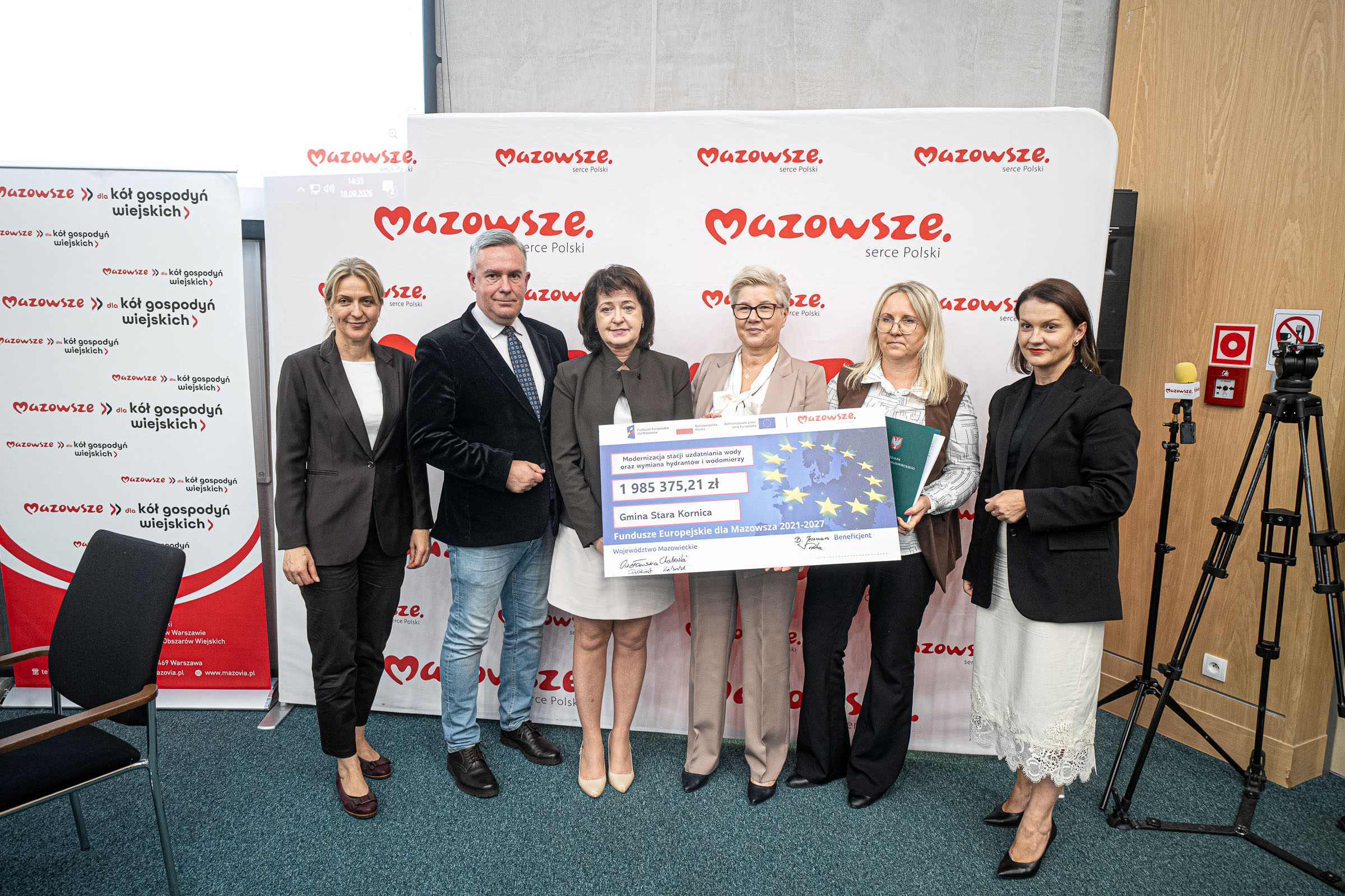 Sześć osób — pięć kobiet i jeden mężczyzna — stoi w rzędzie na tle białego baneru z czerwonym napisem "Mazowsze", ubrani formalnie, trzy z nich trzymają duży symboliczny czek z motywem gwiazd UE, po lewej widoczny roll‑up również z logo "Mazowsze", po prawej statyw z kamerą i ściana z drewnianą okładziną oraz oznaczeniami przeciwpożarowymi.