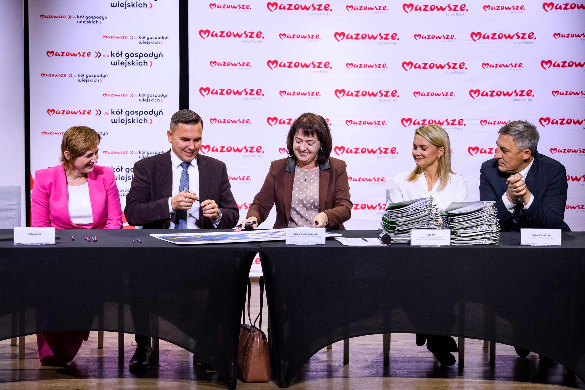 Pięć osób siedzi za długim stołem z czarnym obrusem podpisując dokumenty — od lewej kobieta w różowej marynarce, mężczyzna w ciemnym garniturze, kobieta w brązowej marynarce podpisująca arkusz, kobieta w białej bluzce przy wysokim stosie teczek i mężczyzna w ciemnym garniturze; w tle powtarzający się czerwony napis Mazowsze na białym banerze.