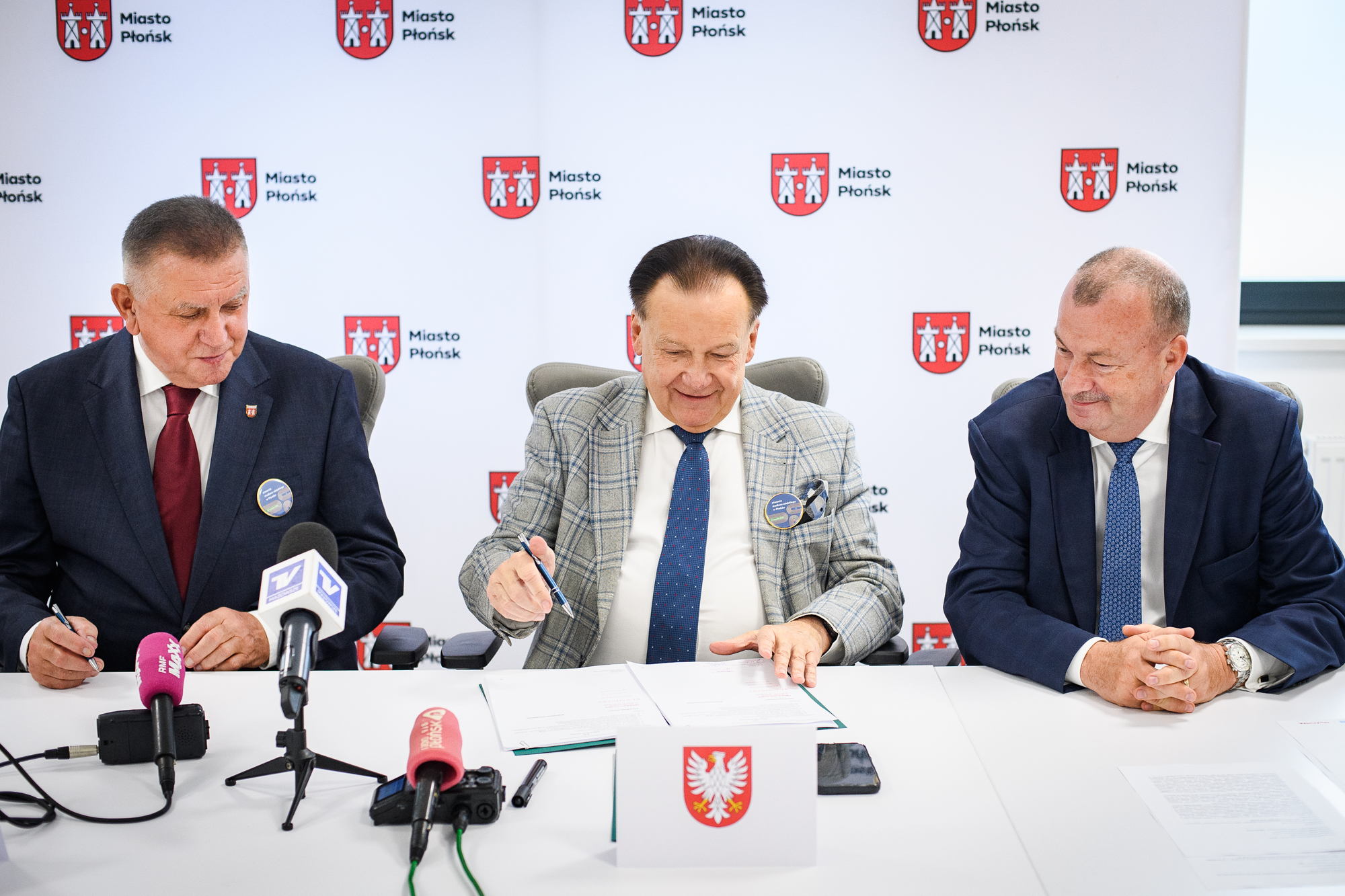 Trzech mężczyzn w garniturach siedzi przy stole i podpisuje dokumenty; w tle baner z powtarzającym się czerwonym herbem i napisem "Miasto Płońsk", na stole widoczne papiery, długopisy, kilka mikrofonów z kolorowymi osłonami oraz mała stojąca kartka z godłem Polski.