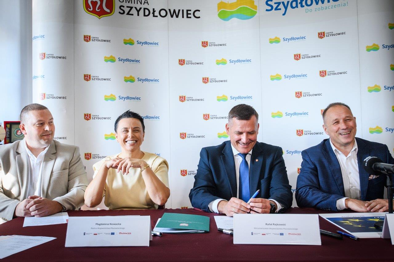 Trzech mężczyzn i kobieta siedzą obok siebie przy bordowym stole z dokumentami, długopisami i tabliczkami imiennymi; kobieta w jasnej bluzce, mężczyźni w marynarkach, a za nimi biały baner z powtarzającymi się logotypami i napisem Gmina Szydłowiec.