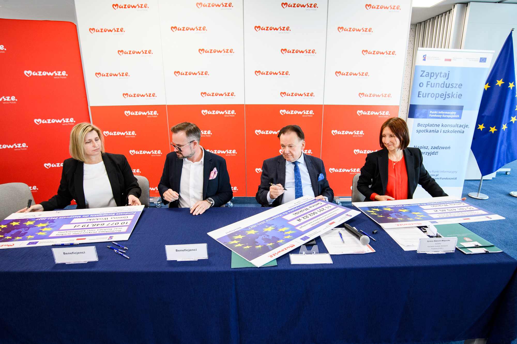 Cztery osoby w formalnych ubraniach siedzą przy długim niebieskim stole i podpisują duże symboliczne czeki i dokumenty, za nimi czerwono-białe panele z powtarzającym się logo „Mazowsze”, po prawej widoczna flaga Unii Europejskiej i stojący baner z napisem „Zapytaj o Fundusze Europejskie”, a na stole leżą tabliczki z napisem „Beneficjenci”, długopisy, mikrofon i materiały informacyjne.
