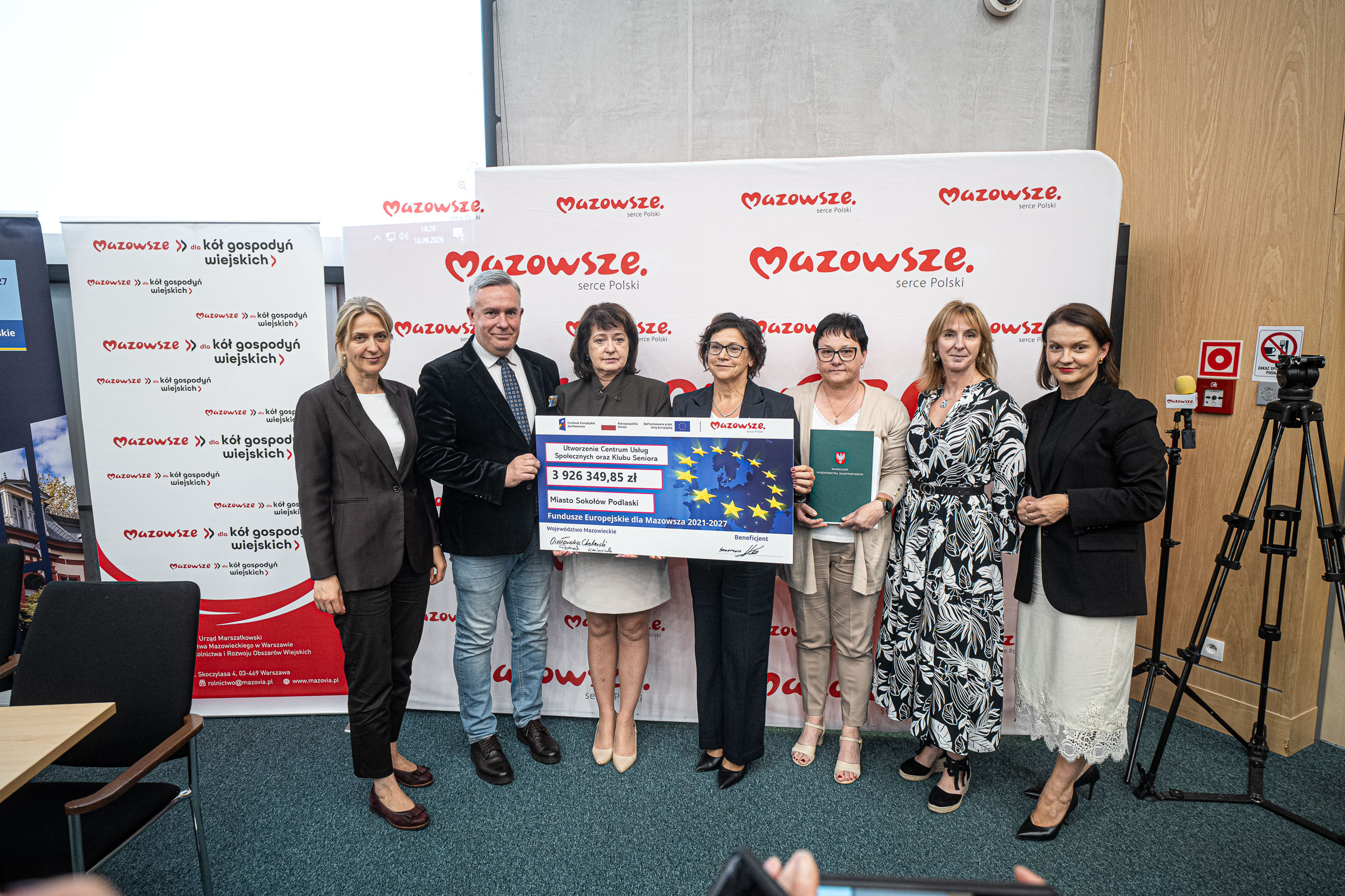 Grupa siedmiu osób w oficjalnym ubiorze stoi w rzędzie na tle białego baneru z powtarzającym się czerwonym logo „Mazowsze serce Polski” i trzyma duży symboliczny czek z emblematem Unii Europejskiej oraz kwotą 3 926 349,85 zł, obok widoczne roll-upy z napisem „Mazowsze”, kamera na statywie i elementy wyposażenia ściennego w sali konferencyjnej.