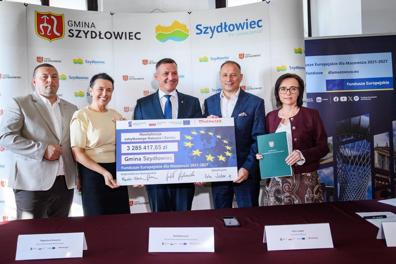 Pięć osób stoi za stołem konferencyjnym przed banerami z logo "Gmina Szydłowiec" i "Szydłowiec", trzymając duży czek z napisem "Rewitalizacja zabytkowego Ratusza i Zamku" na kwotę 3 285 417,65 zł z motywem gwiazd UE, a po prawej kobieta trzyma zielony folder z herbem.