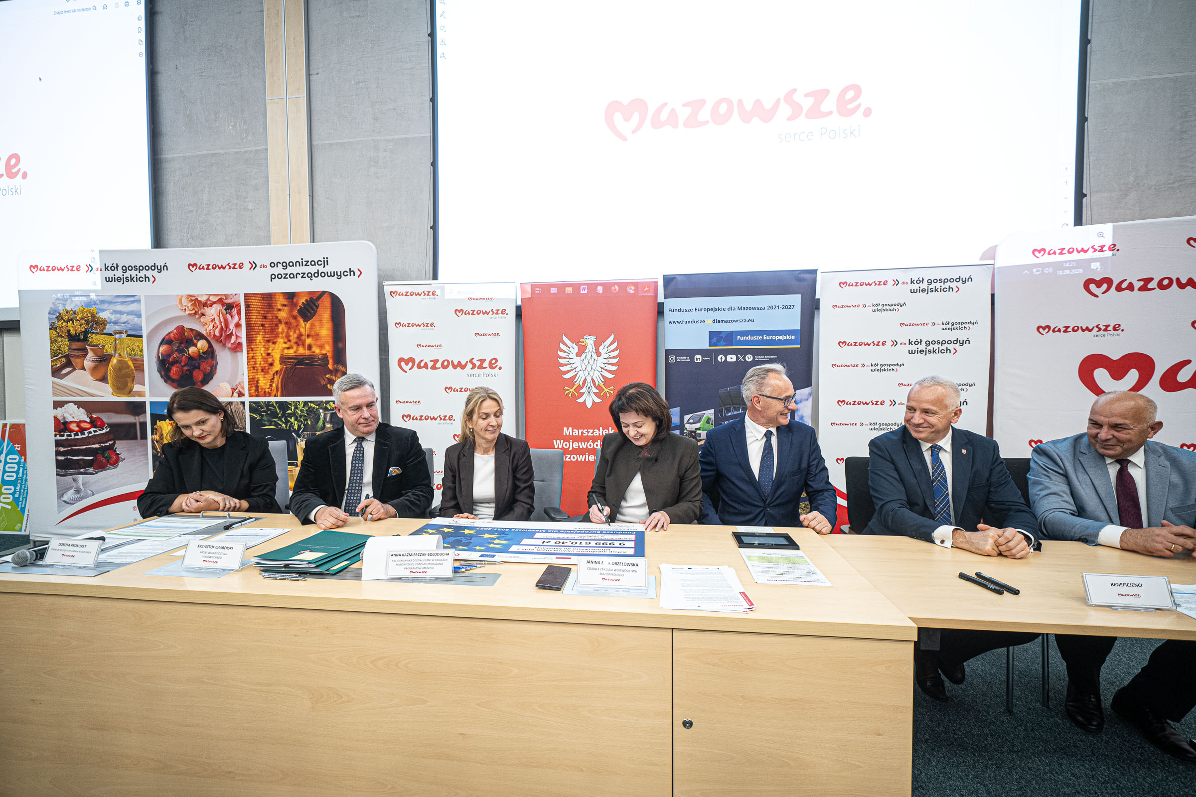 Siedem osób w formalnych strojach siedzi przy długim jasnym stole, podpisując lub przeglądając dokumenty; na stole leżą teczki, formularze, mikrofon i tabliczki z nazwiskami, a w tle duże ekrany i banery z logo „Mazowsze”, herbem białego orła oraz grafikami promocyjnymi.