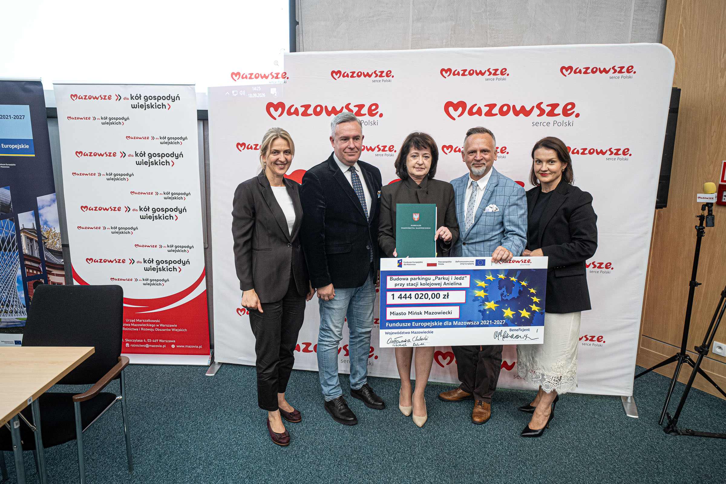 Pięcioro dorosłych stoi obok siebie na tle białego baneru z powtarzającym się czerwonym napisem "Mazowsze", a w środku osoba trzyma zieloną teczkę i duży czek z kwotą 1 444 020,00 zł; po obu stronach widać stojące roll-upy promocyjne, po lewej stół i krzesło, po prawej statyw z mikrofonem.