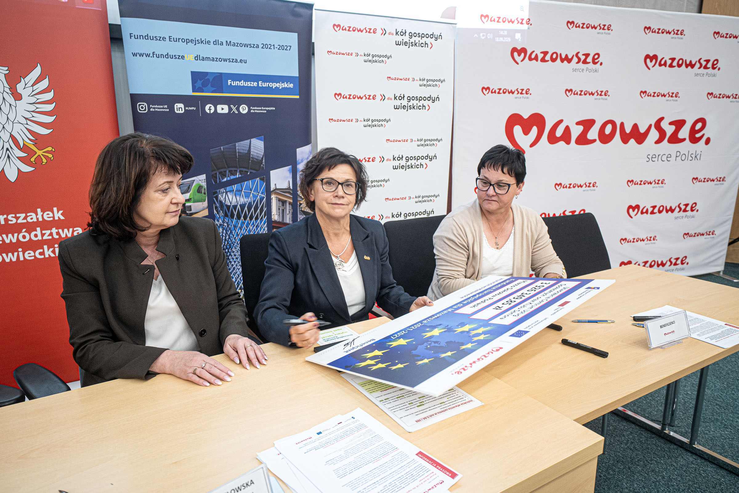 Trzy kobiety siedzą przy stole przeglądające i podpisujące duży plakat z gwiazdami UE; w tle banery z logo „Mazowsze” i napisem „Fundusze Europejskie dla Mazowsza 2021–2027”, na stole dokumenty, długopisy i identyfikator „Beneficenci”.