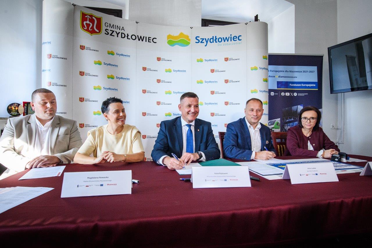 Pięć osób siedzi za stołem z bordowym obrusem przed składanym banerem z napisem i logotypami „Gmina Szydłowiec” i „Szydłowiec”, na stole leżą dokumenty oraz tabliczki z nazwiskami, a centralny mężczyzna w granatowym garniturze trzyma długopis przy papierach; po prawej widać stojący ekran telewizora.